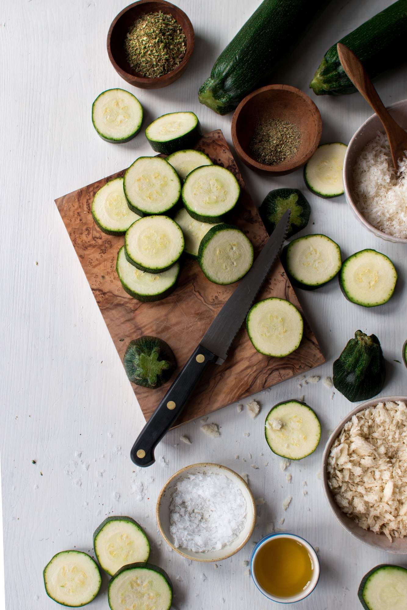 Ingredients for crispy baked zucchini including sliced zucchini, panko breadcrumbs, parmesan cheese, spices, and olive oil.