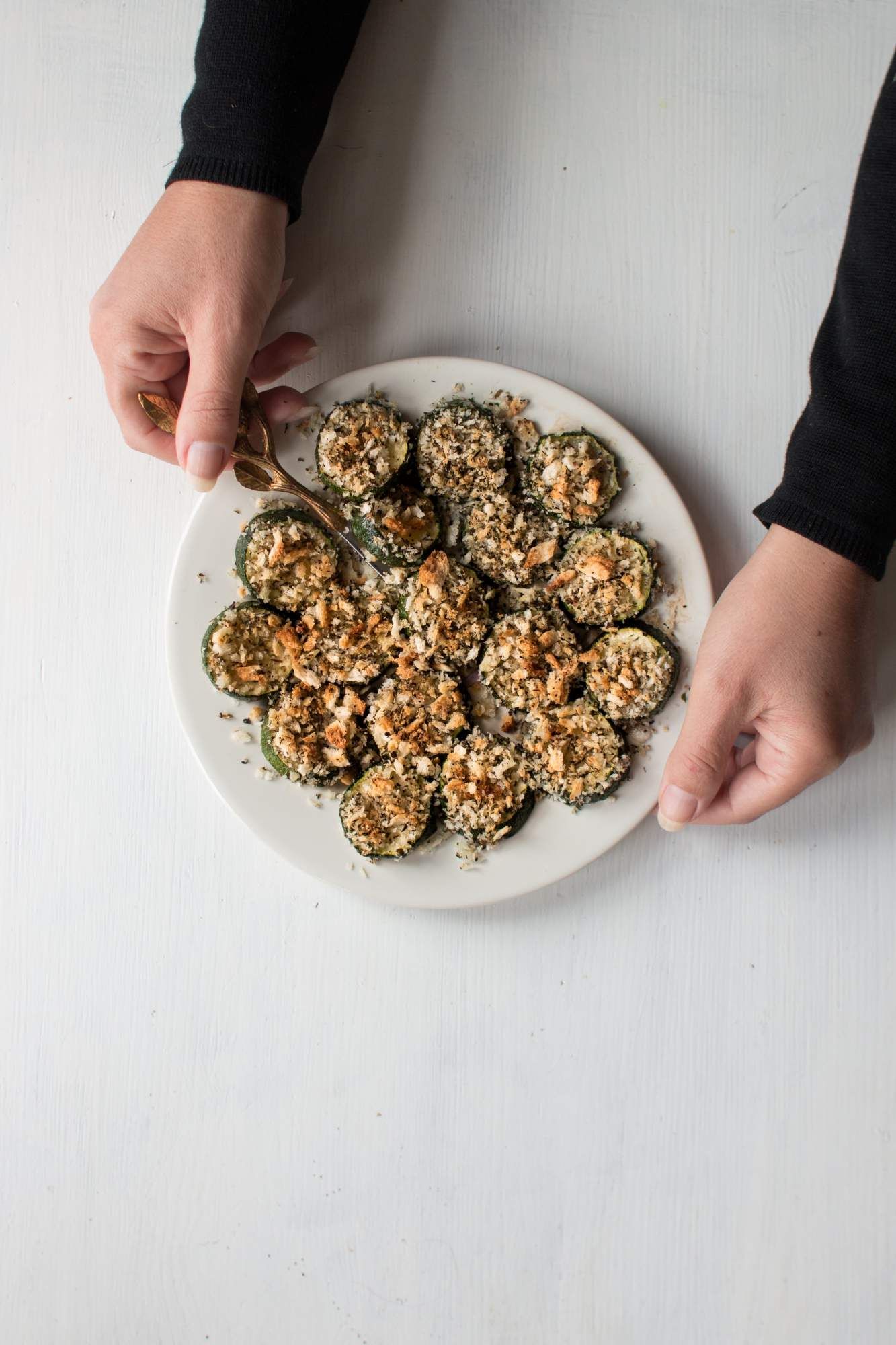 Baked zucchini with crispy parmesan breadcrumbs on a plate with two hands holding it.