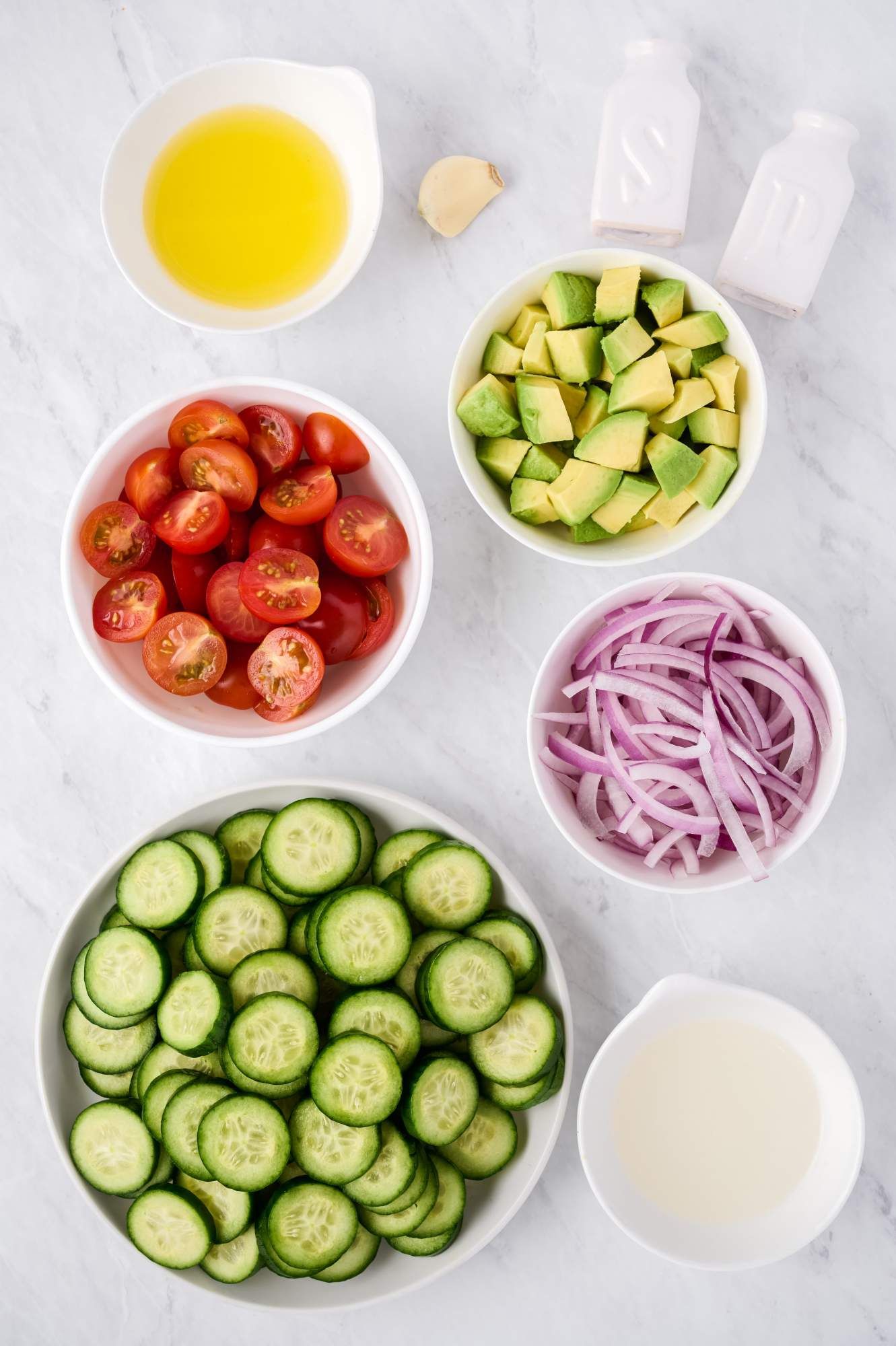 ingredients for cucumber salad including sliced Persian cucumbers, tomatoes, red onions, avocado, olive oil, garlic, and white vinegar.