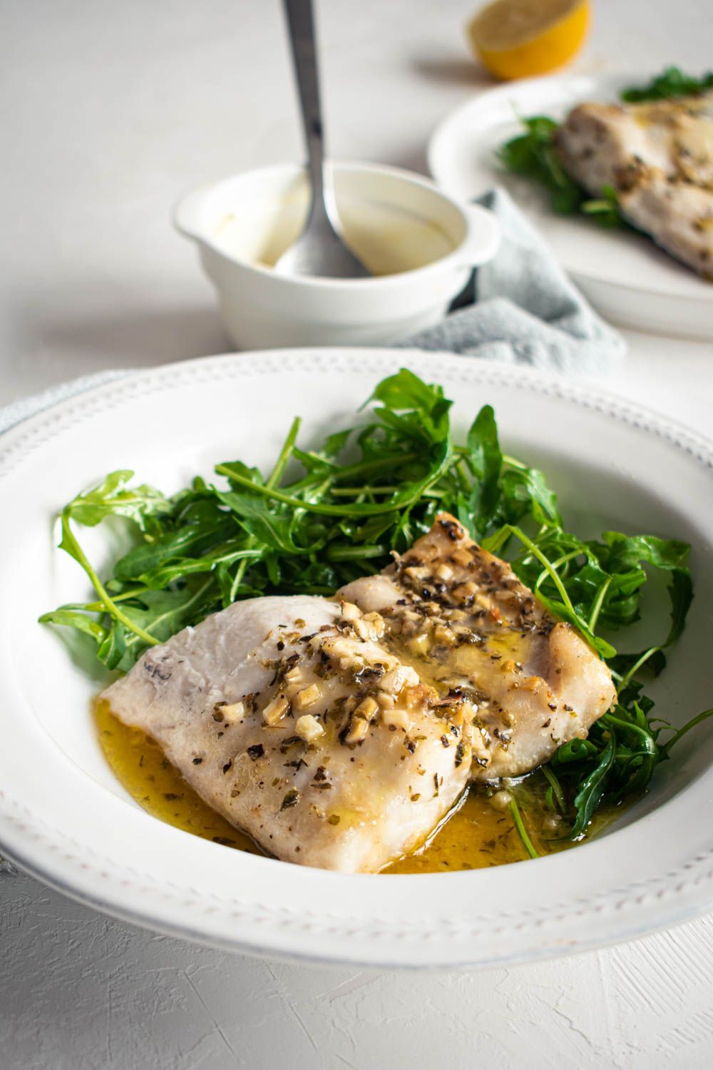 Lemon baked cod with garlic and herbs on a white plate with butter sauce and green salad.