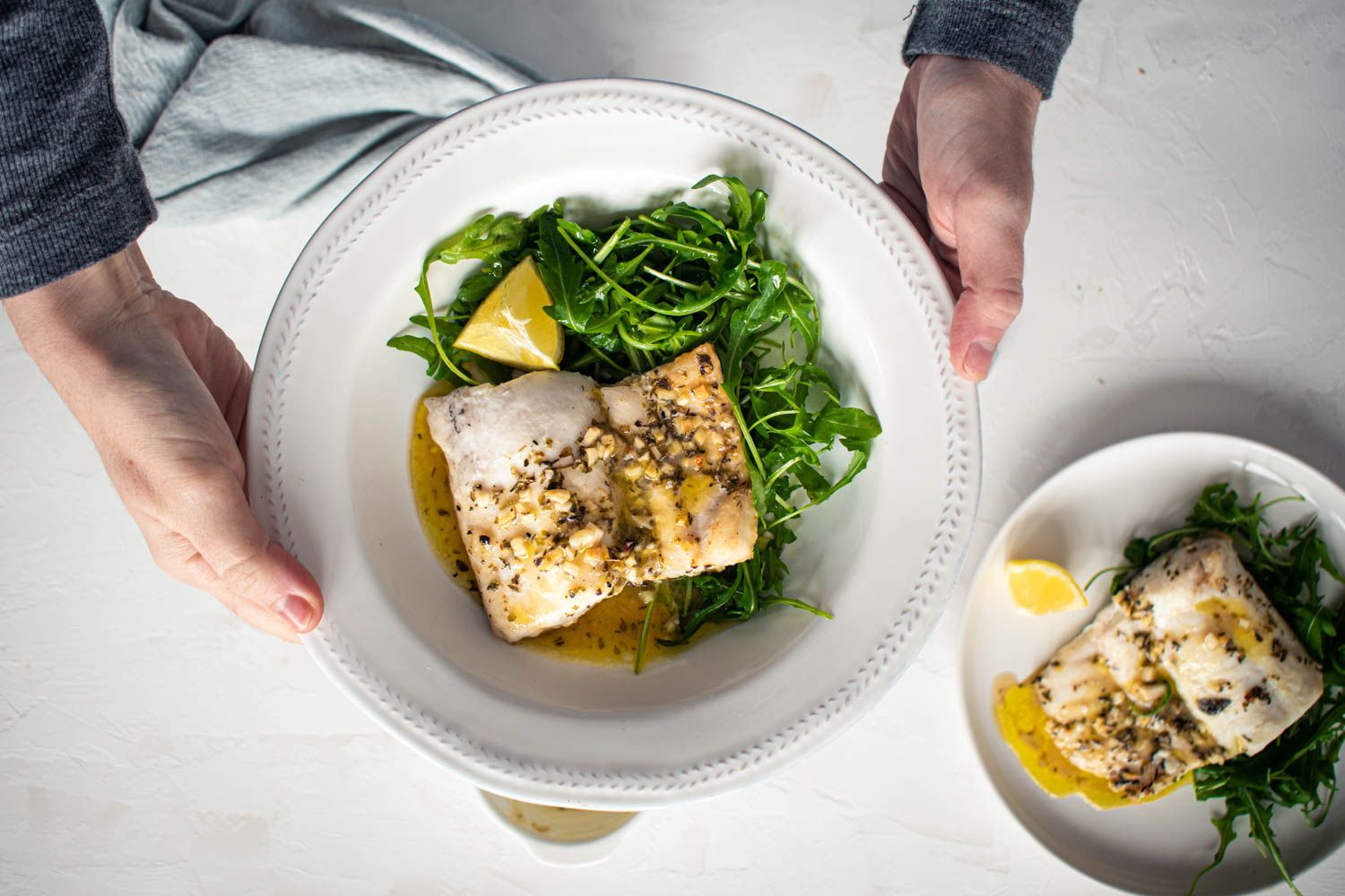 Cod filets that are baked with garlic, butter, lemon, and seasoning on a plate with green salad and a blue napkin.