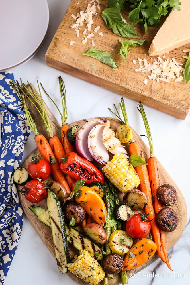 Grilling vegetables piled onto a platter with fresh parsley including corn, tomatoes, peppers, zucchini, and carrots.