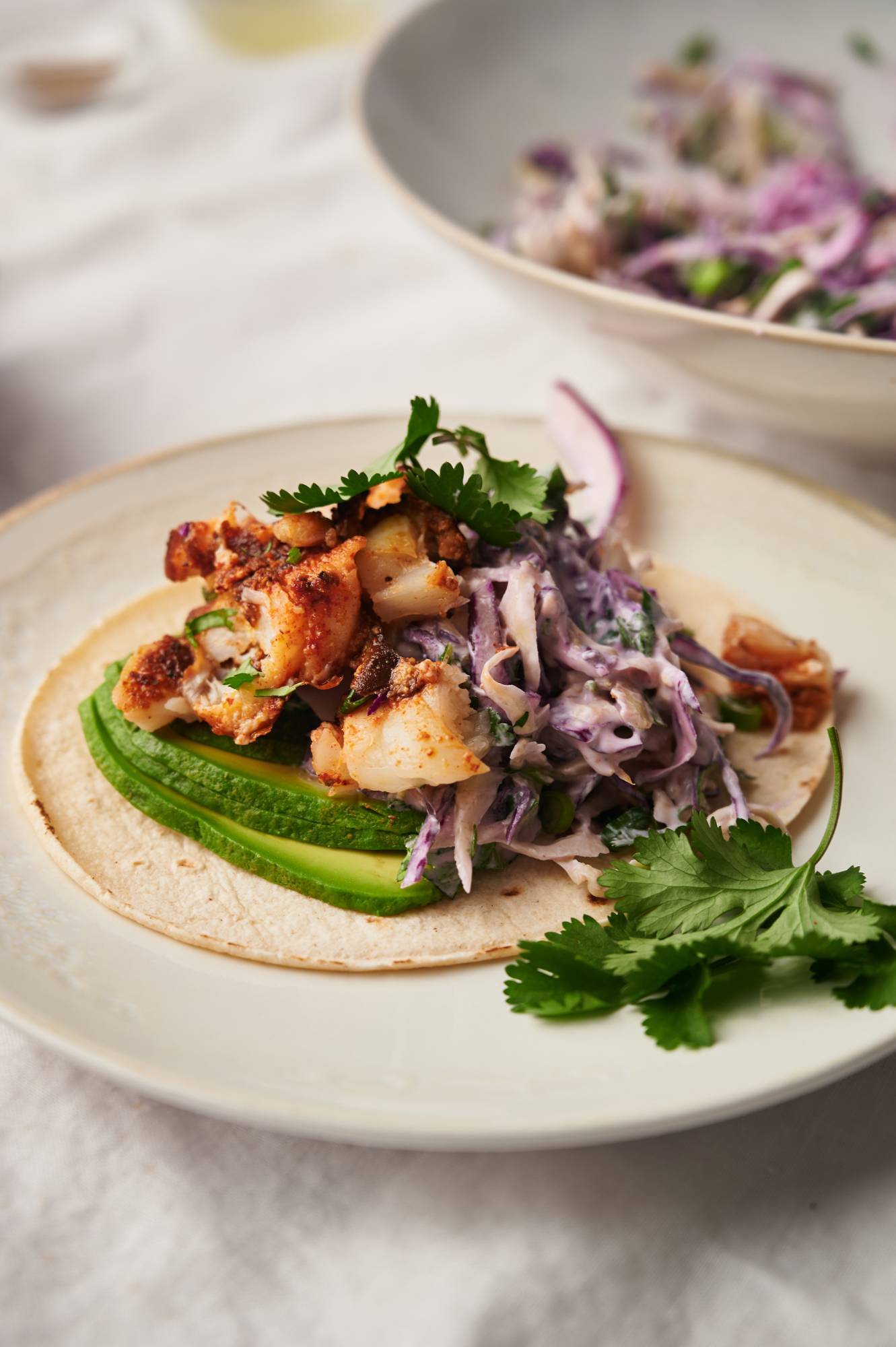 Fish taco with slaw, seasoned fish, and avocado on a corn tortilla with cilantro.