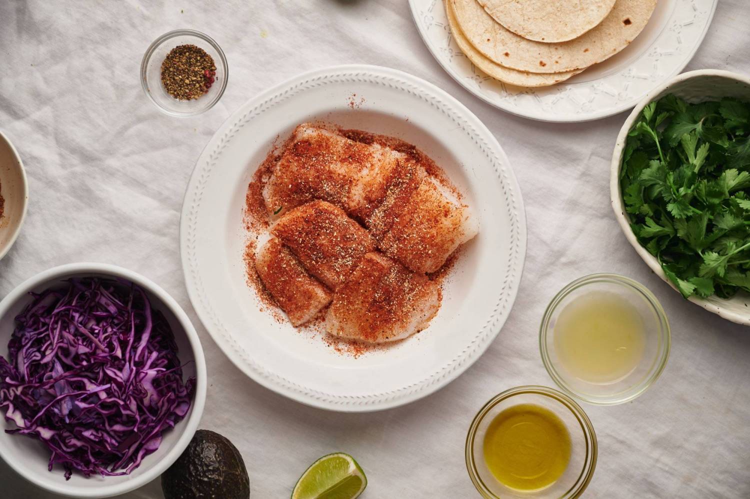 White fish covered in taco seasoning in a bowl with cabbage, cilantro, tortillas, and lime juice on the side.