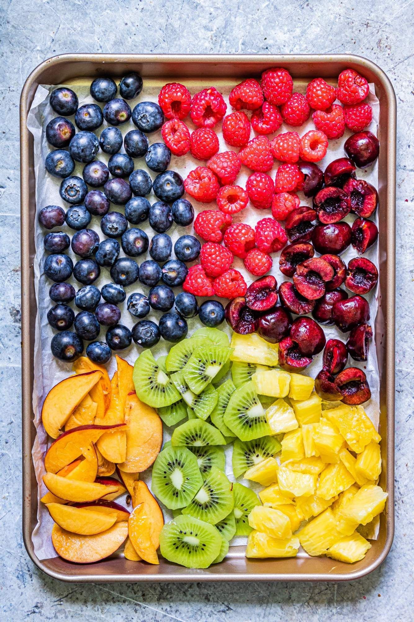 Homemade fruit popsicle ingredients including berries, pineapple, cherries, and peaches on a baking sheet.