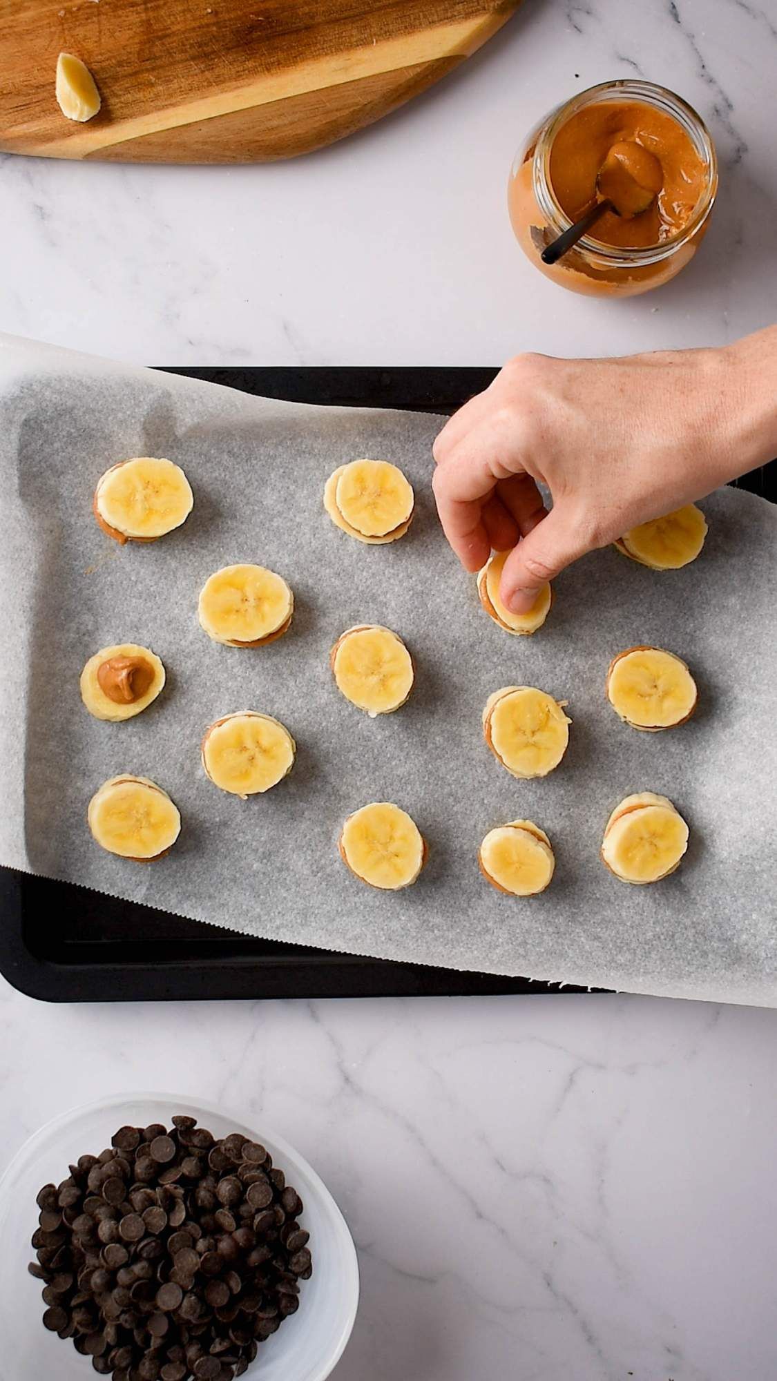 Banana slices topped with peanut butter on a baking sheet with chocolate chips on the side,