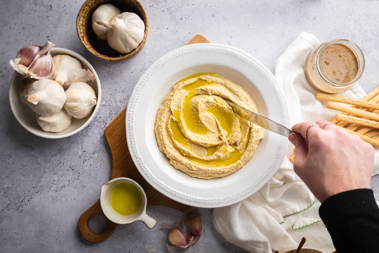 Homemade hummus with roasted garlic and olive oil in a bowl with garlic bulbs and breadsticks on the side.