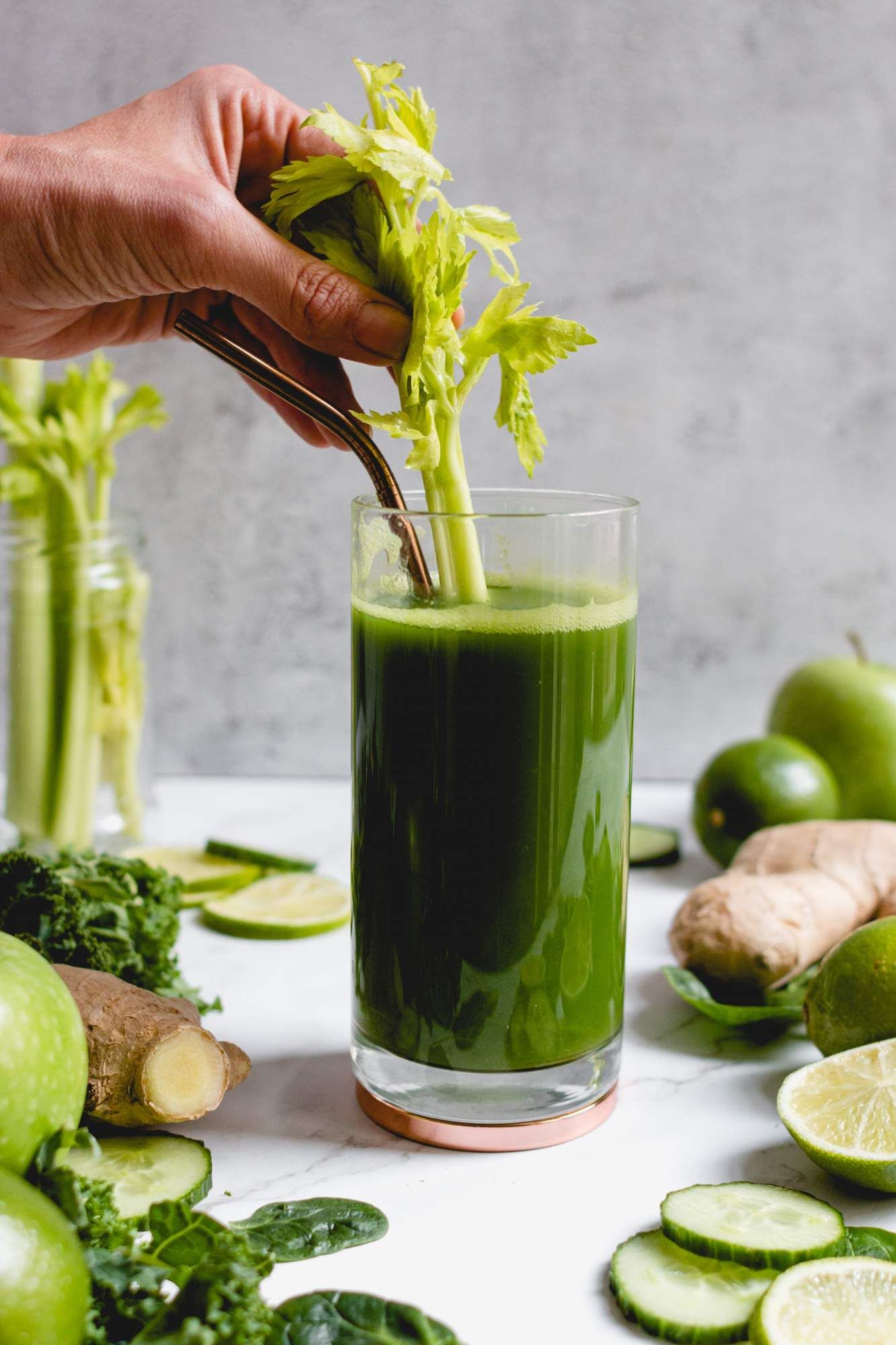 Blender green juice  in a glass with cucumbers, celery, apples, and ginger in the side.