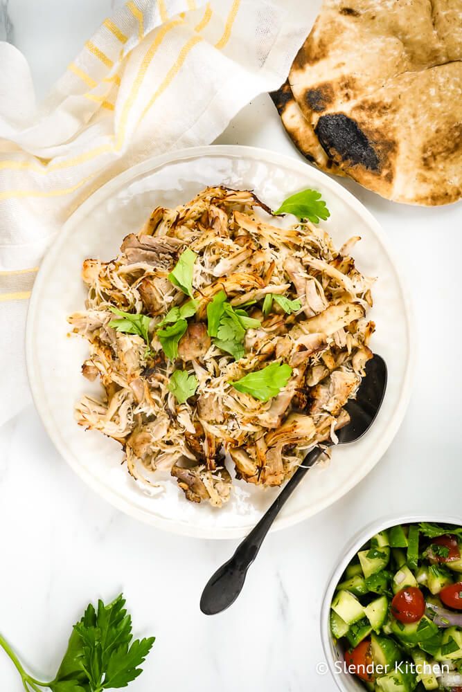 Greek shredded chicken on a white plate with parsley, tomato salad, and pita bread.