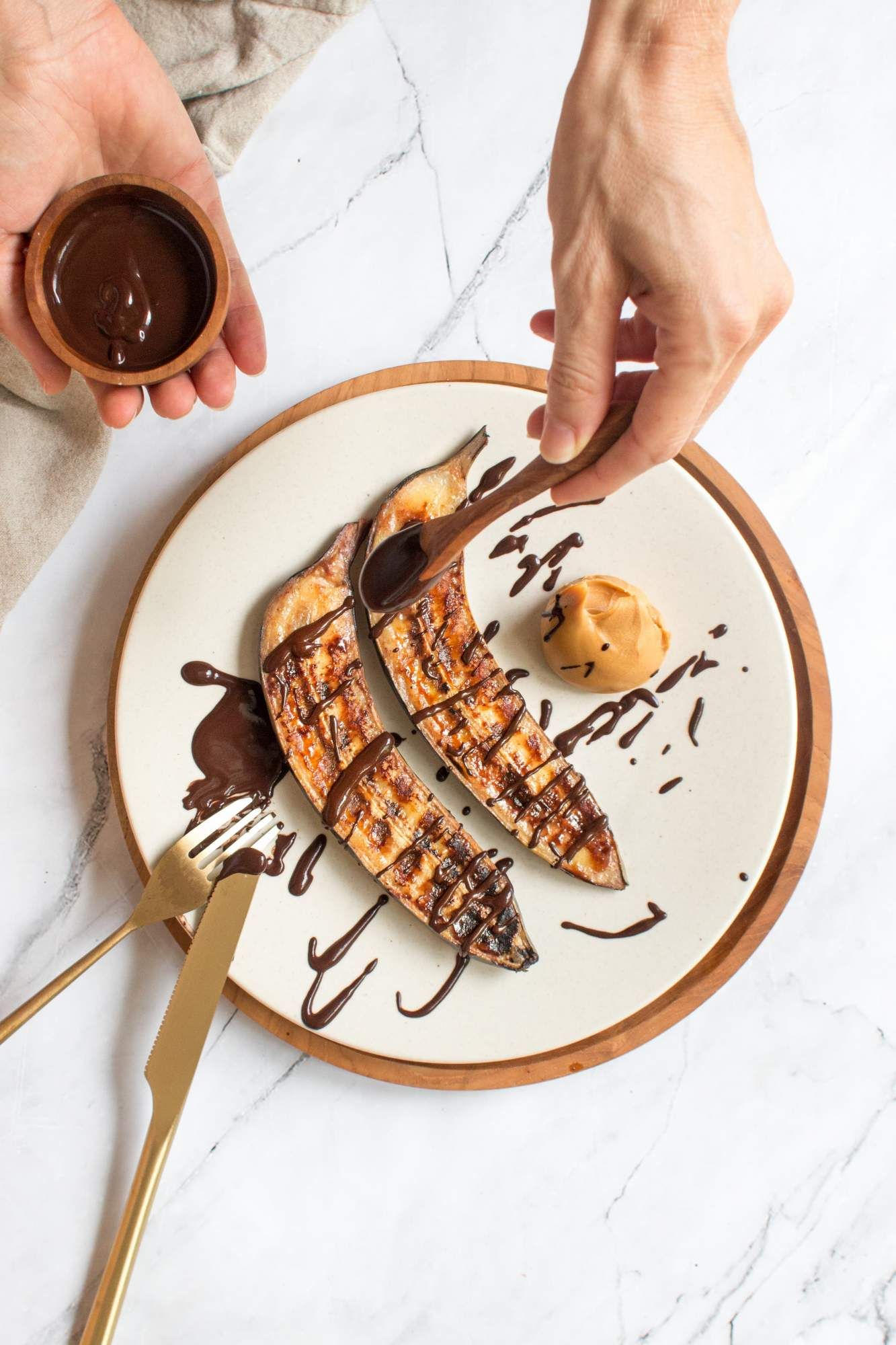 Grilled bananas drizzled with peanut butter and chocolate on a plate with a hand adding chocolate.