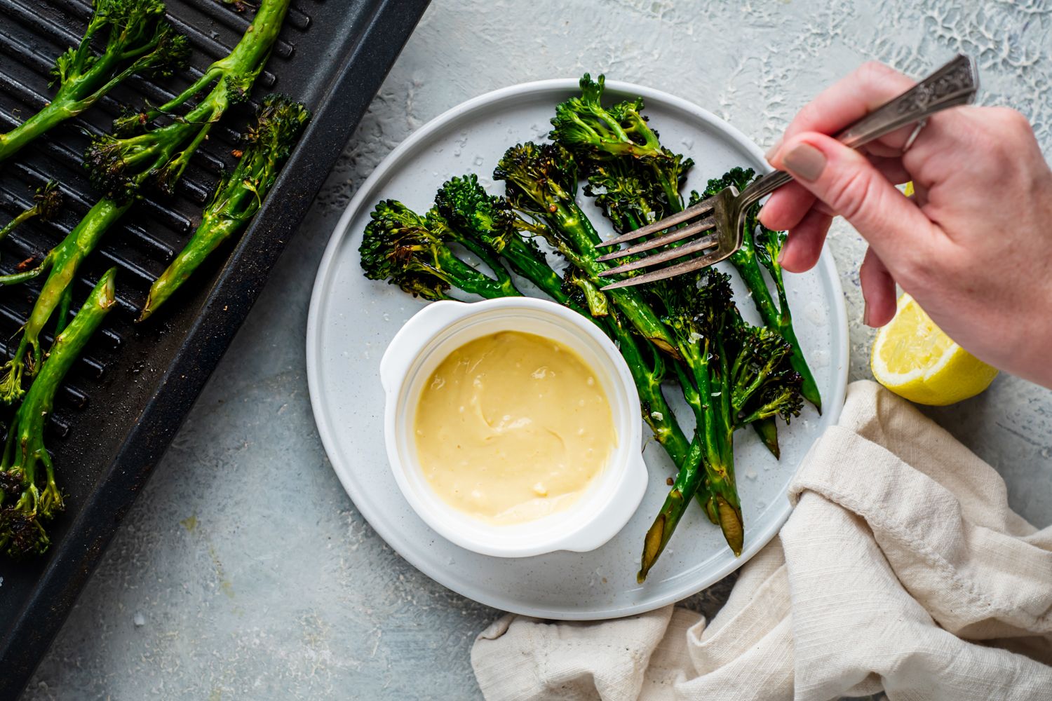 Crispy grilled broccolini on a plate and on a grill pan with lemon, garlic aioli, and black pepper.