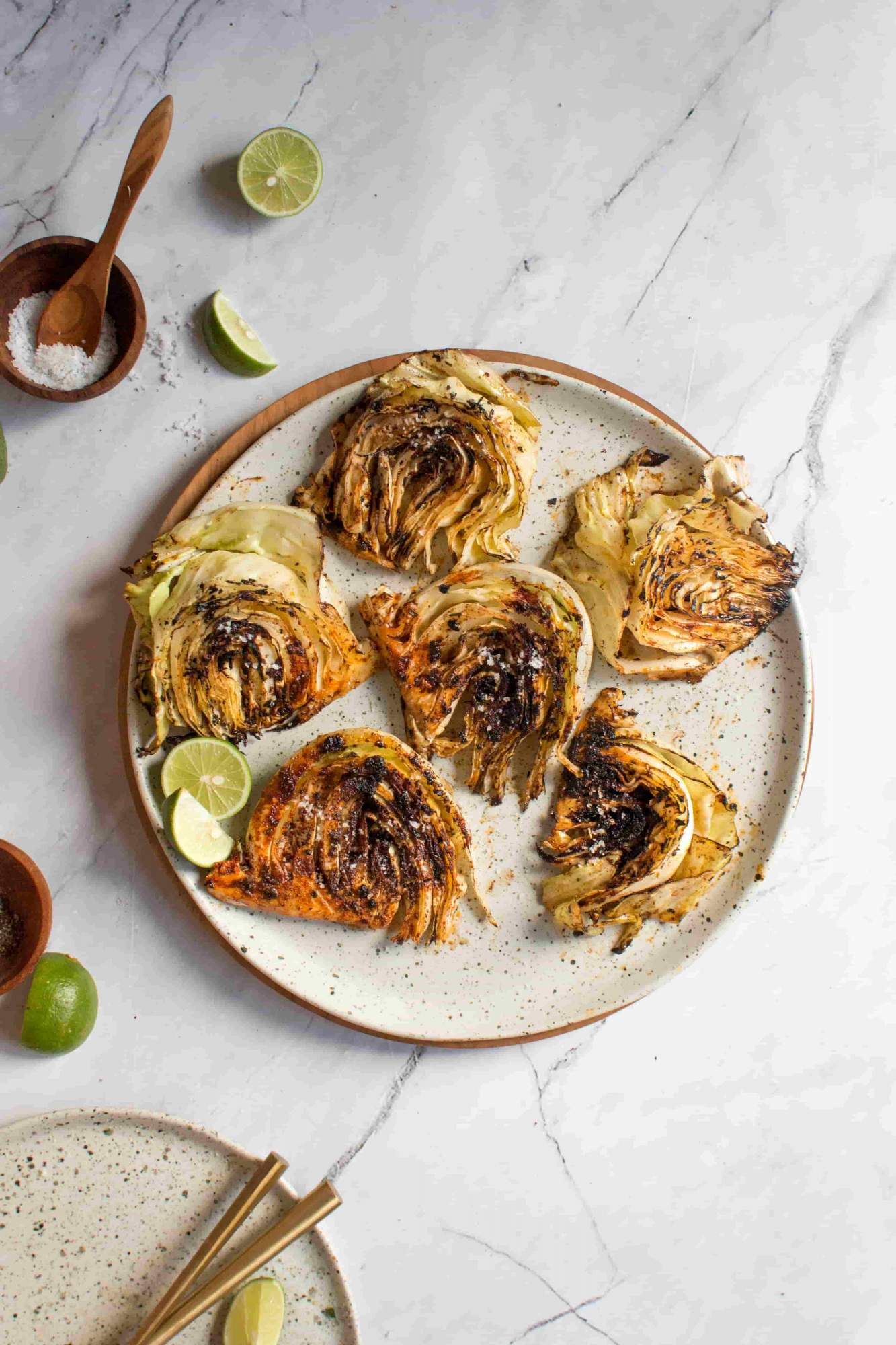 Grilled cabbage steaks on a plate with flaky salt, limes, and spices. 