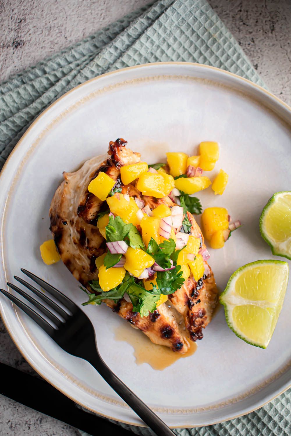 Marinated grilled chicken served with mango salsa on a plate with fresh limes on the side.