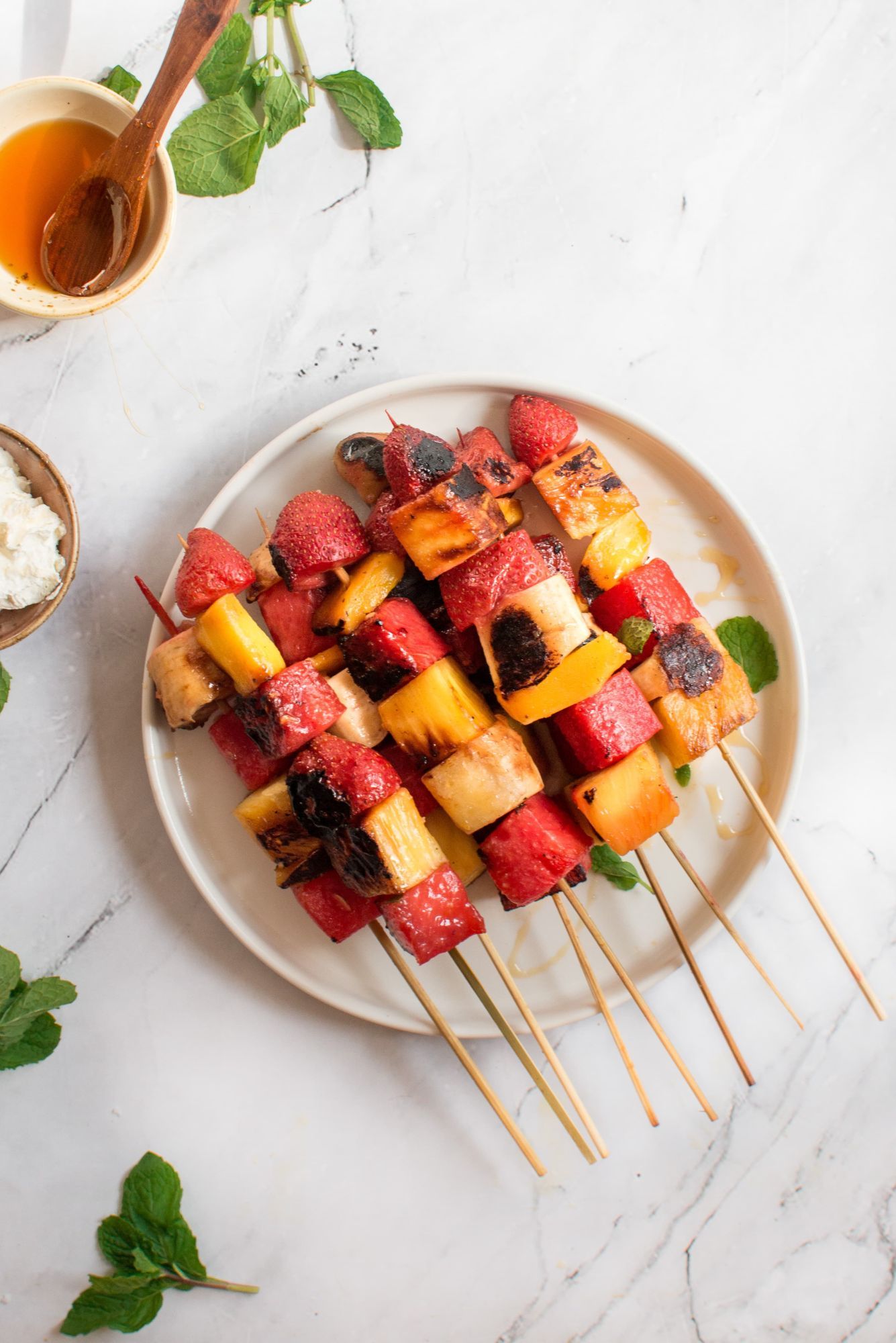 Fruit kabobs with grilled fruit including banana, watermelon, strawberries on a plate with honey and mint,