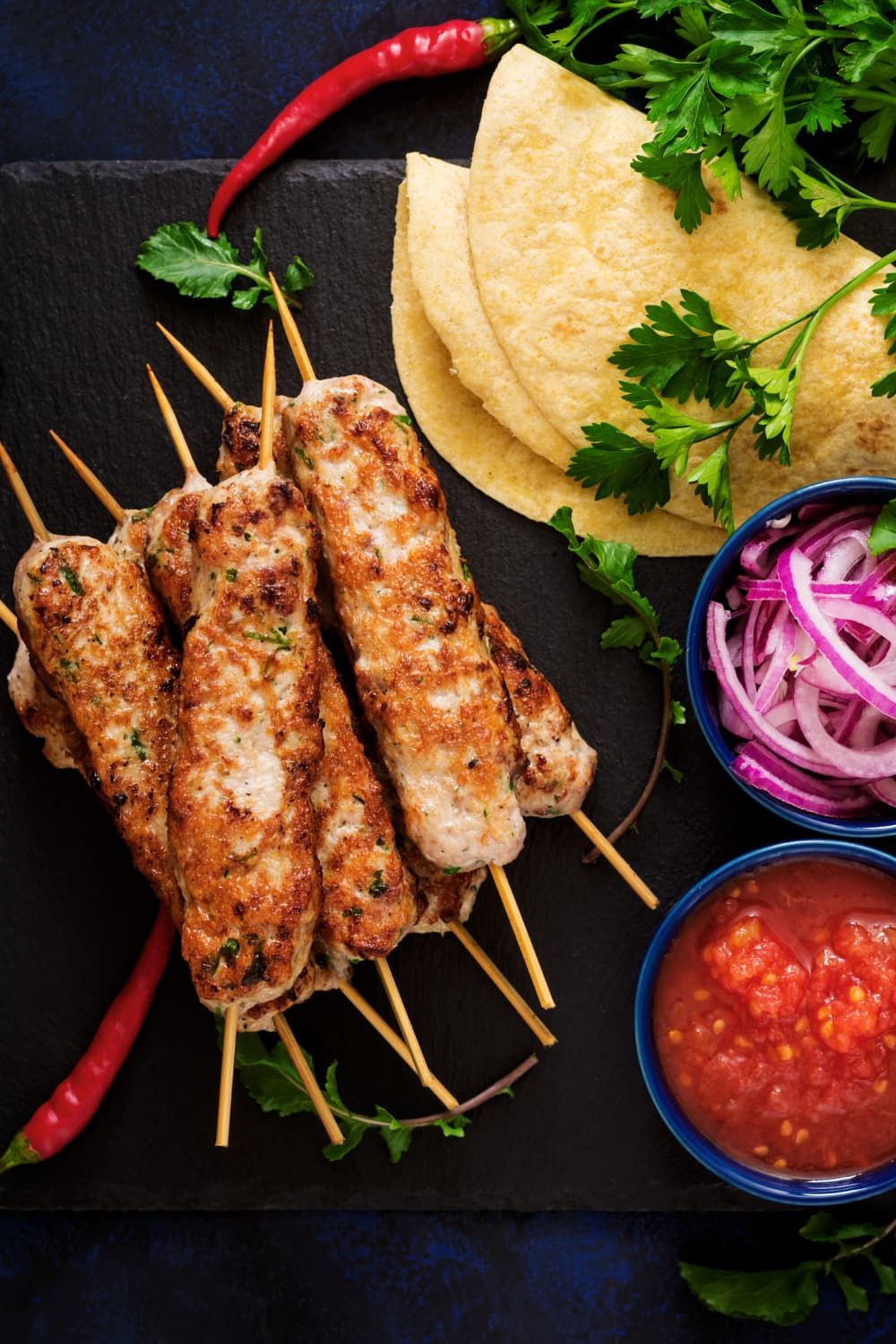 Lula kabobs made with ground turkey on a board with tortillas, red onions, tomatoes, and a slate board.