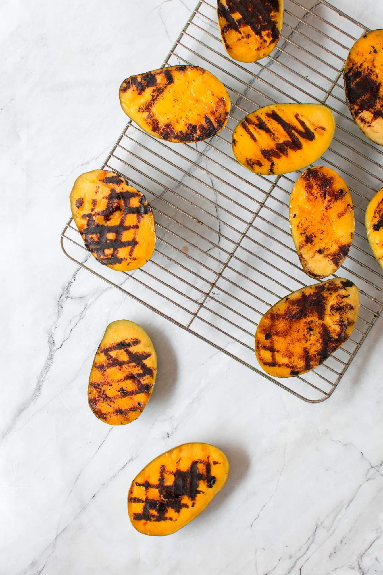 Grilled mangoes sliced in half with grill marks on a wire rack and marble cutting board.