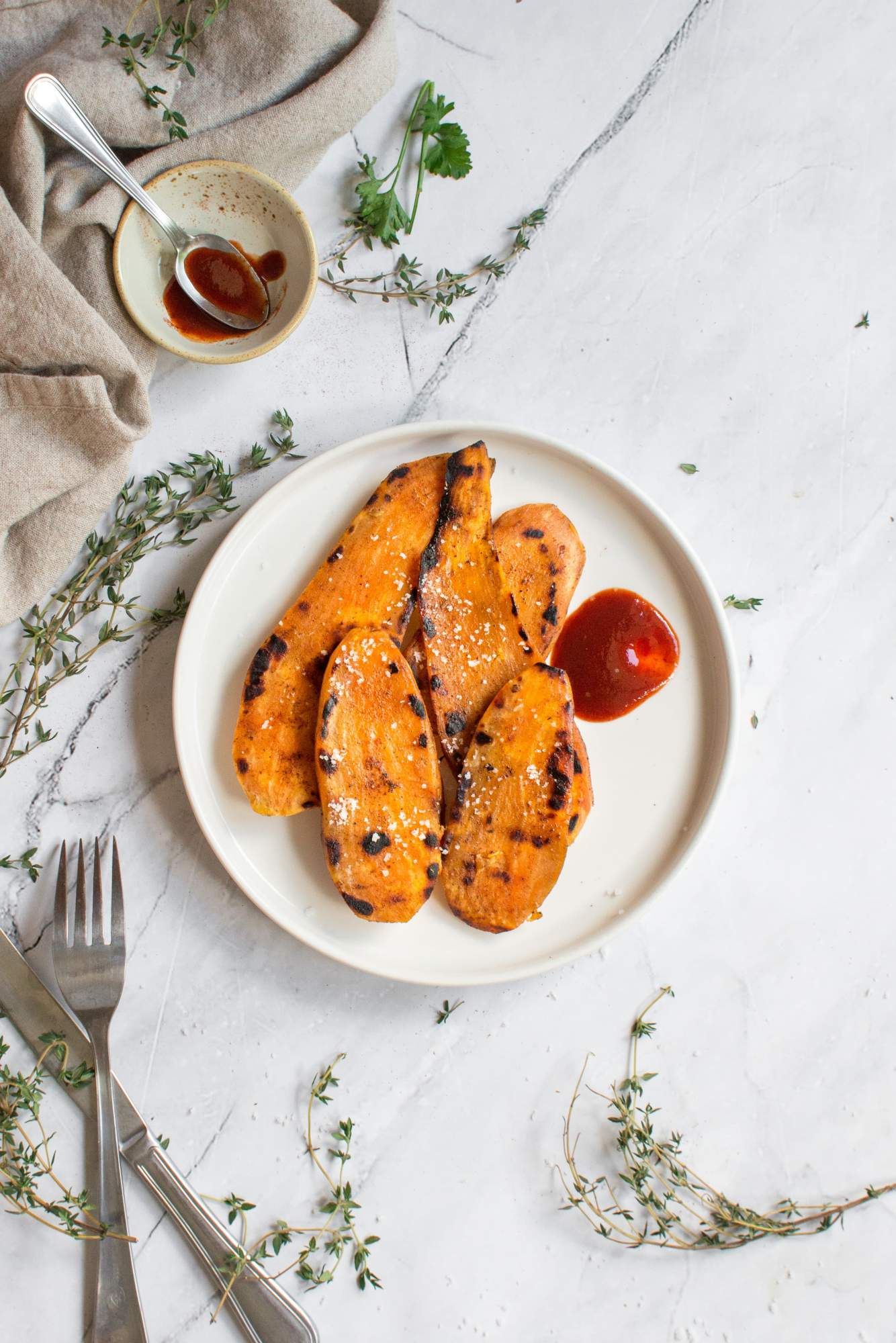 Sweet potatoes that have been grilled on a plate with fresh thyme and barbecue sauce.