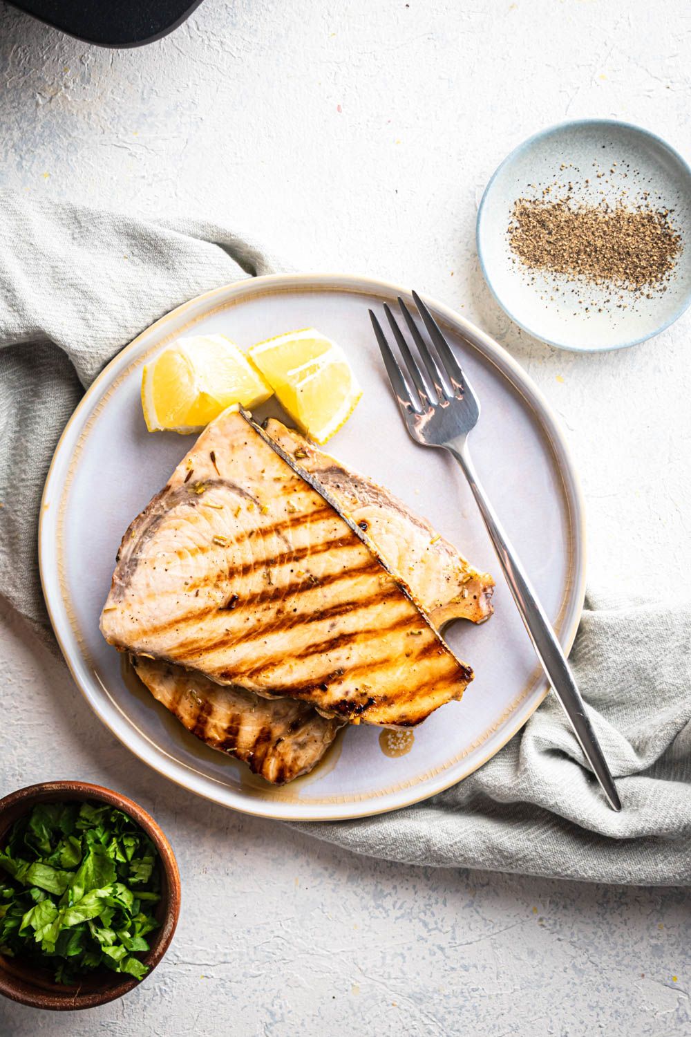 Swordfish that was grilled with soy sauce, lemon, olive oil, garlic, and spices served on a plate with a grill pan on the side.