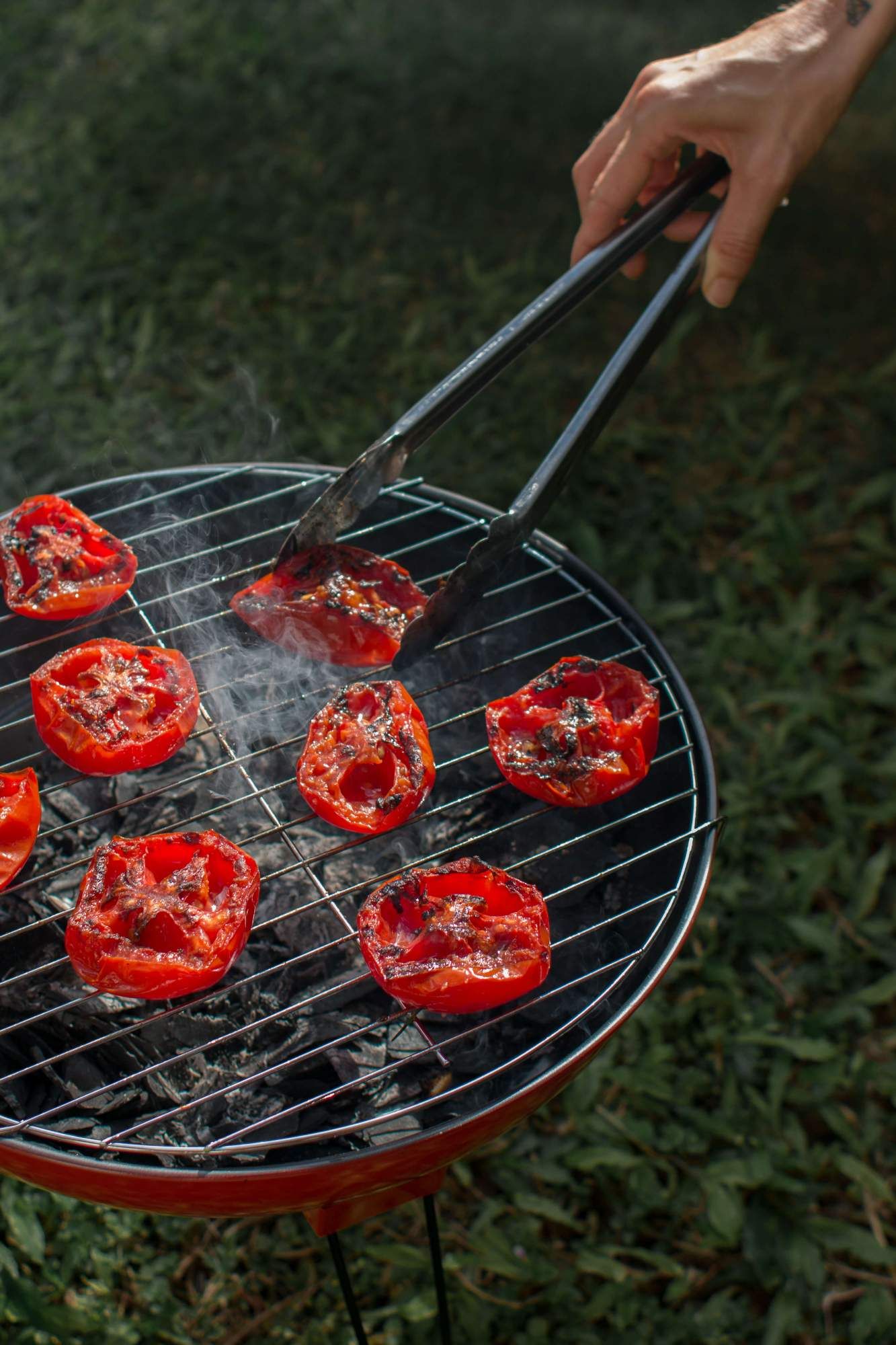 Grilling tomatoes on a charcoal grill and flipping them with tongs.