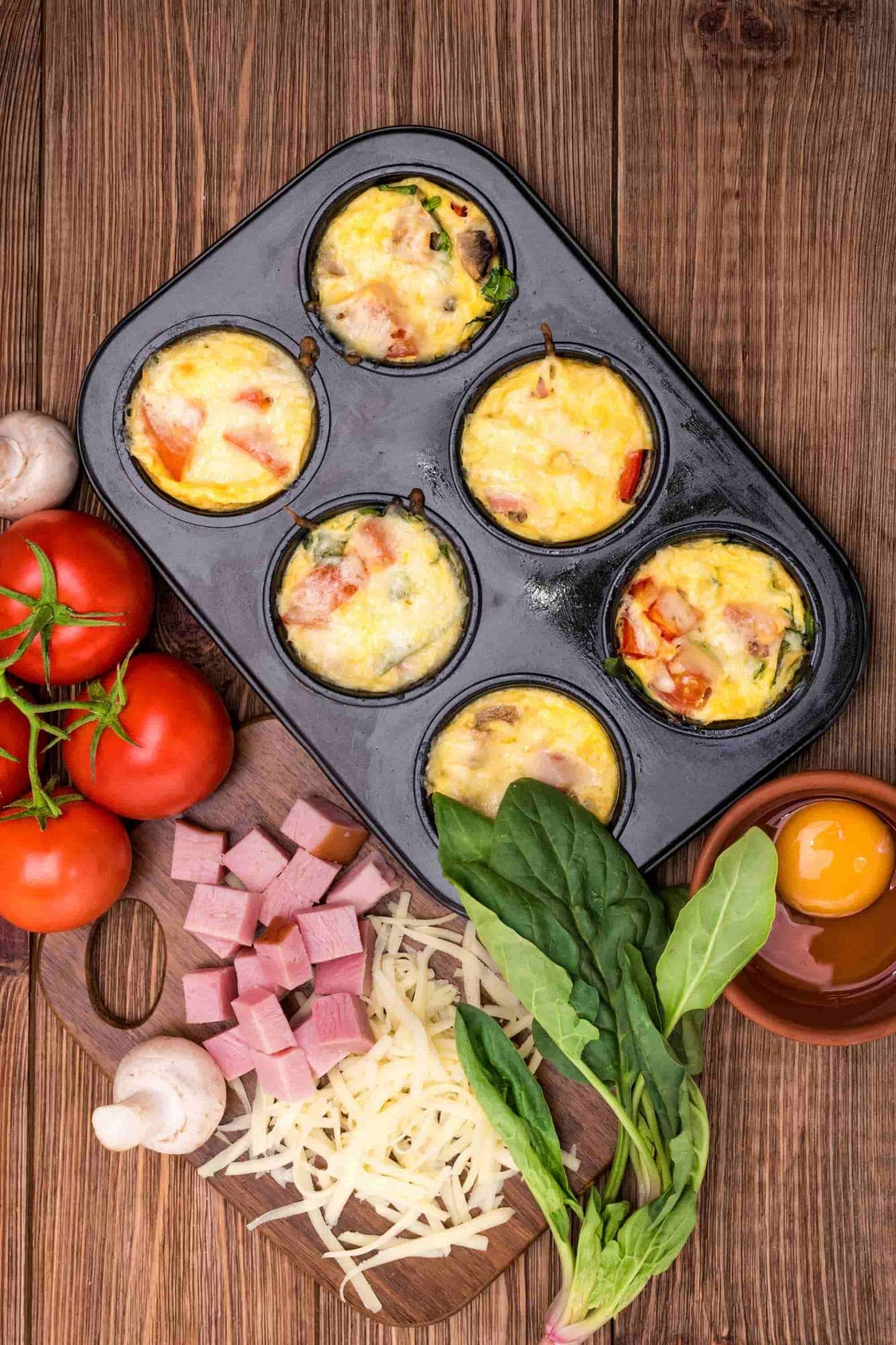 Egg muffins with ham, cheese, tomatoes, and spinach in a muffin tin and with ingredients on the side. 