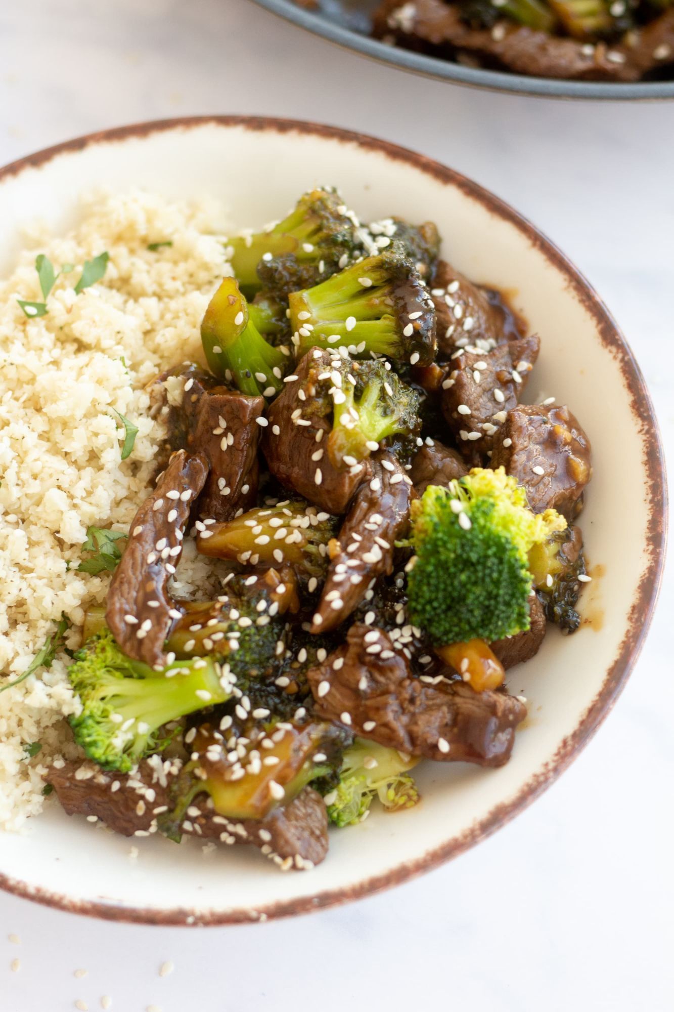 Broccoli and beef stir fry in a bowl with with white rice and sesame seeds in a bowl.