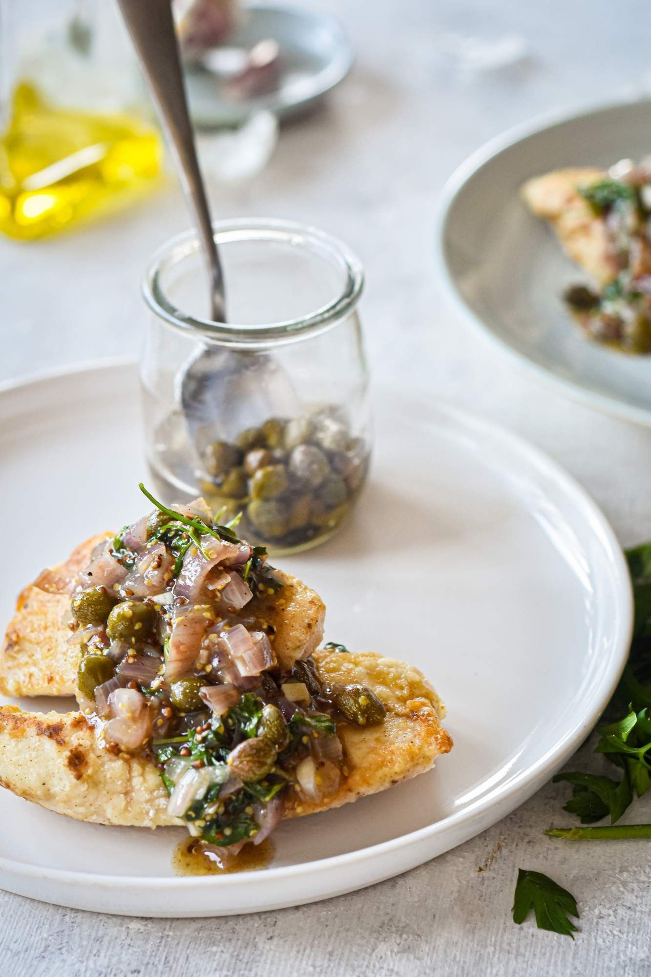 Chicken in piccata sauce with parsley, garlic, lemon, and shallots on a plate with capers in a jar on the side.