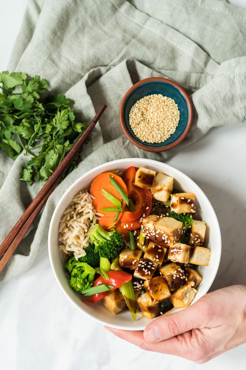 Hoisin tofu with brown rice, broccoli, carrots, celery, and red bell peppers in a bowl with chopsticks and sesame seeds on the side.