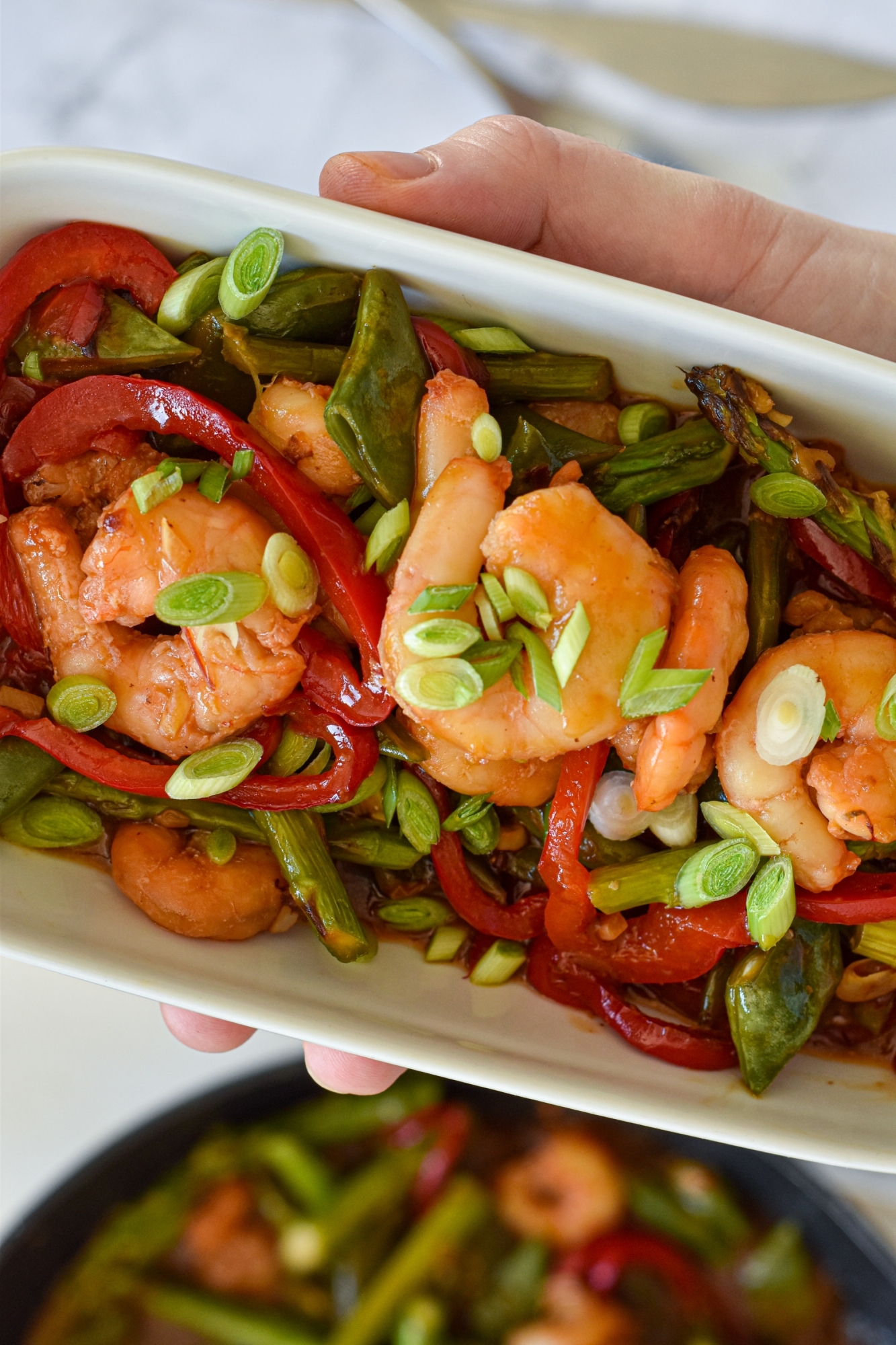 Garlic honey shrimp stir fry in a white dish with snap peas, red bell peppers, asparagus, and green onions.