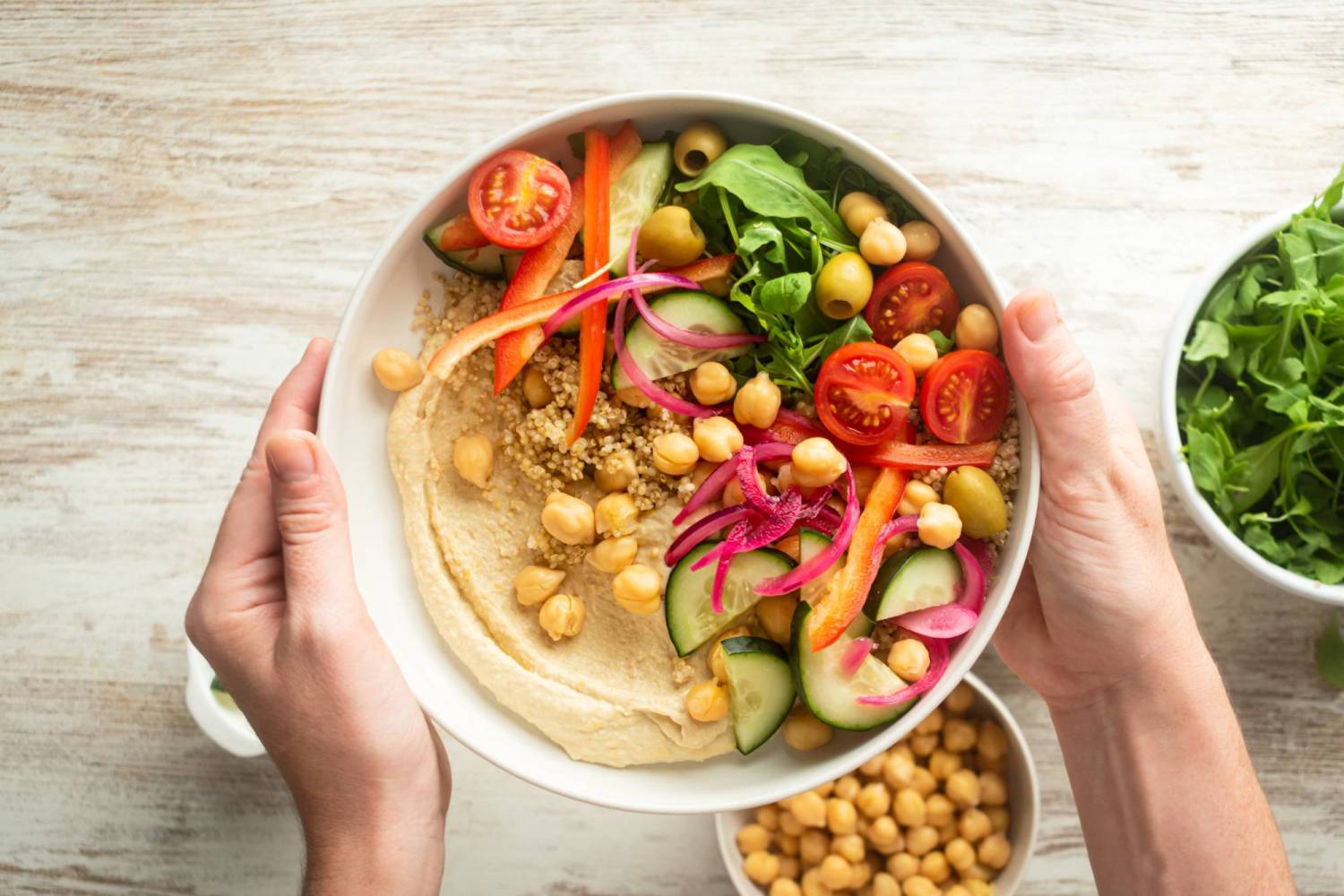 Grain bowl with hummus, greens, red peppers, tomatoes, cherry tomatoes, quinoa, and olives.