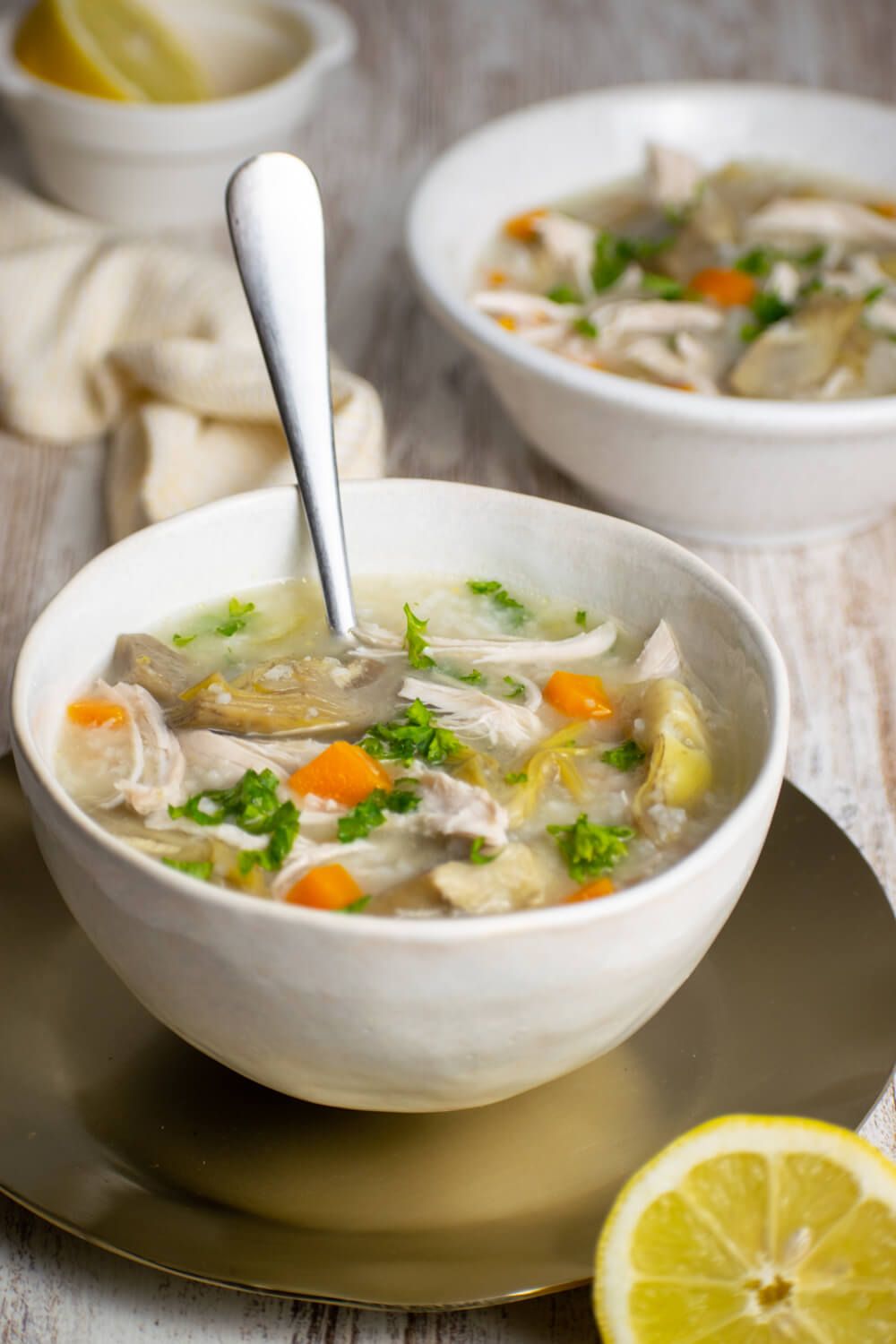 Chicken soup with lemon and artichoke in two white bowls with lemon on the side.