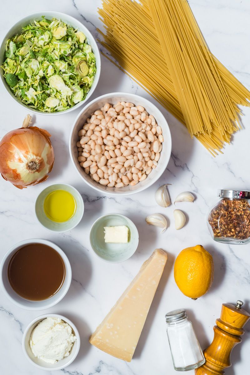 Ingredients for Brussel sprout pasta including shredded sprouts, white beans, cheese, garlic, lemon, and dried pasta.