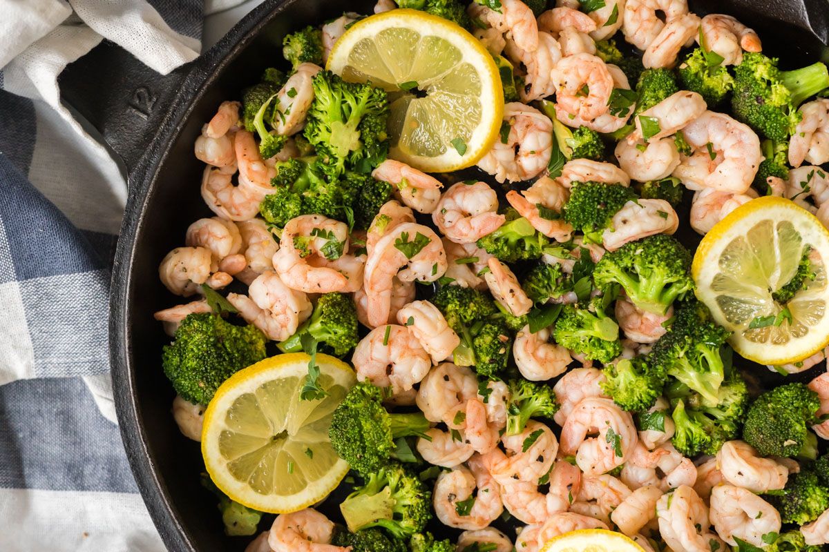 Lemon pepper shrimp served with broccoli and garlic in a skillet with lemon slices and parsley.