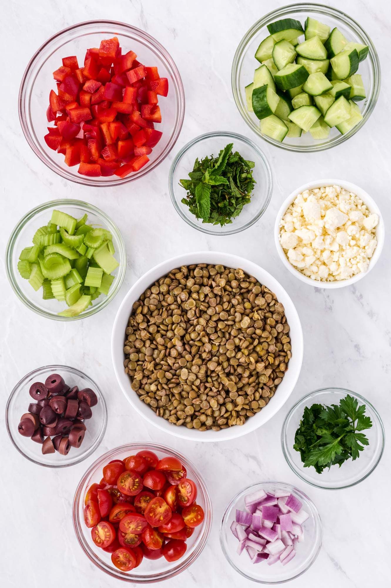 Ingredients for lentil salad including cooked lentils, cucumbers, olives, feta, fresh herbs, and tomatoes.