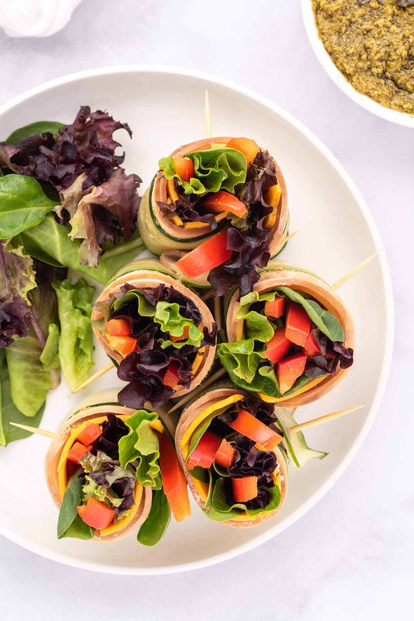 Turkey pesto low carb roll ups in slices of cucumber with greens, red bell pepper, and cheddar cheese.