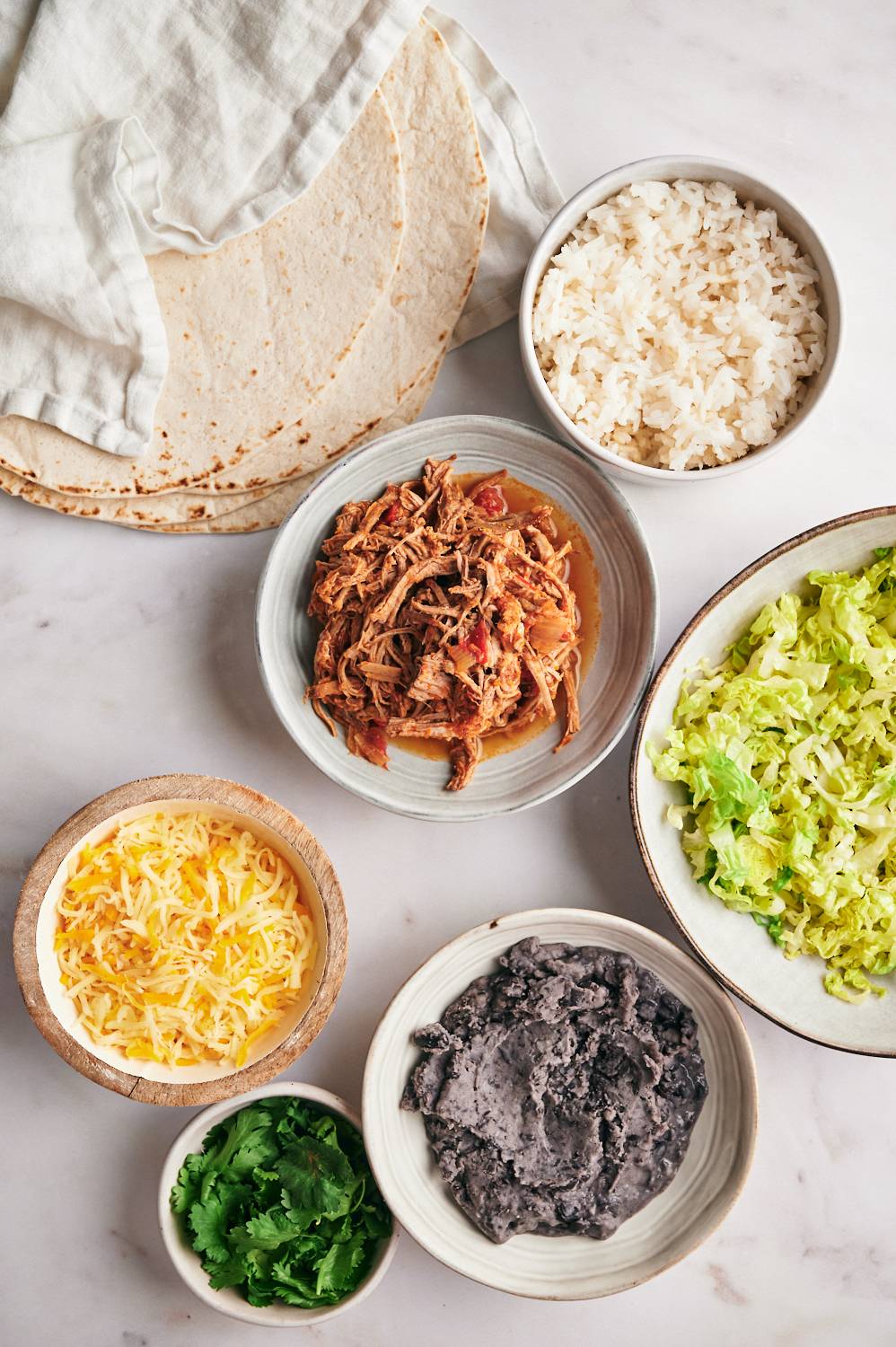 Ingredients for machaca burritos including shredded machaca beef, refried beans, cheese, rice, cilantro, lettuce, and tortillas.