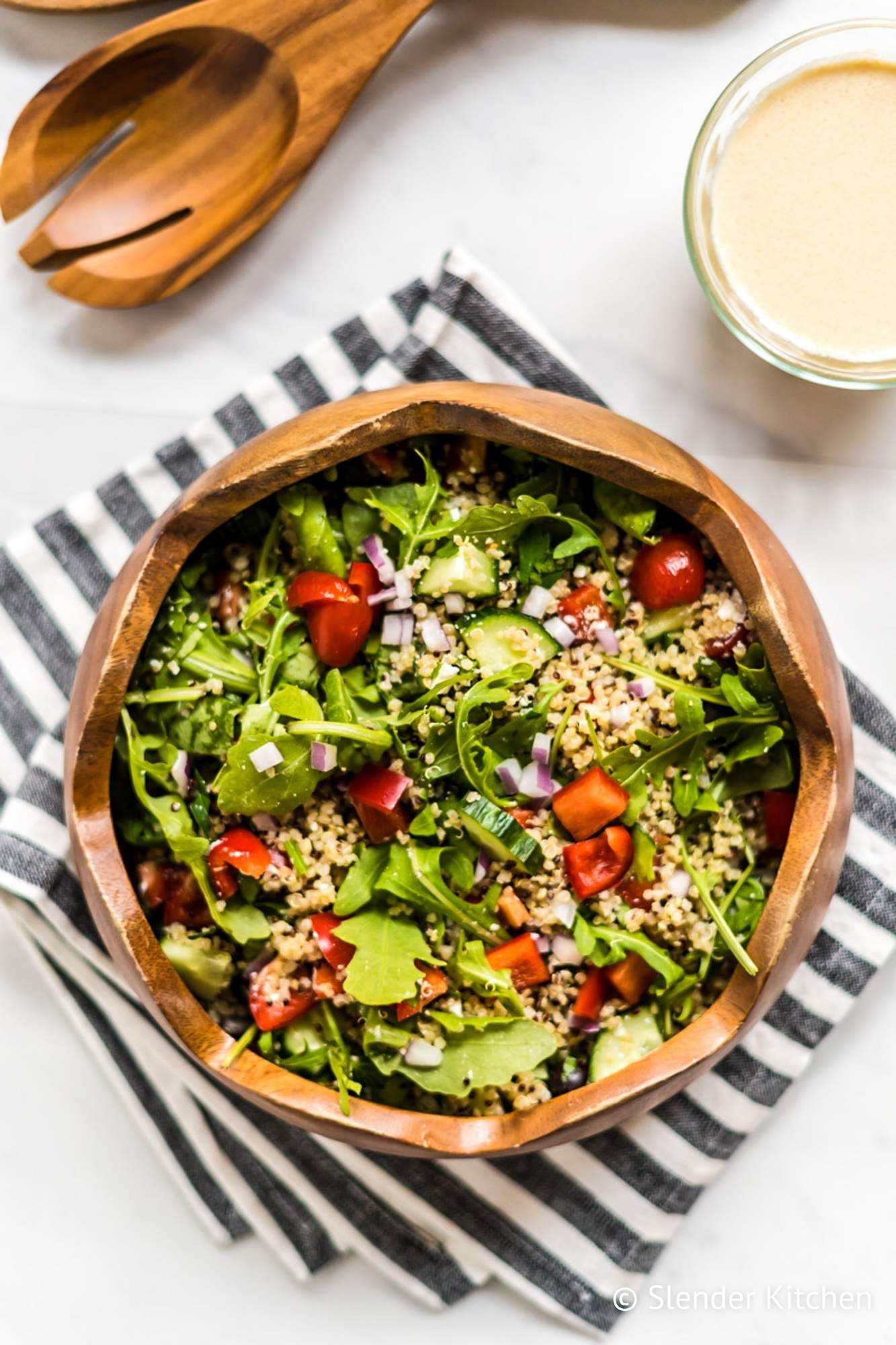 Quinoa salad bowl with Mediterranean flavors including arugula, tomatoes, feta cheese, and black olives in a wooden bowl.