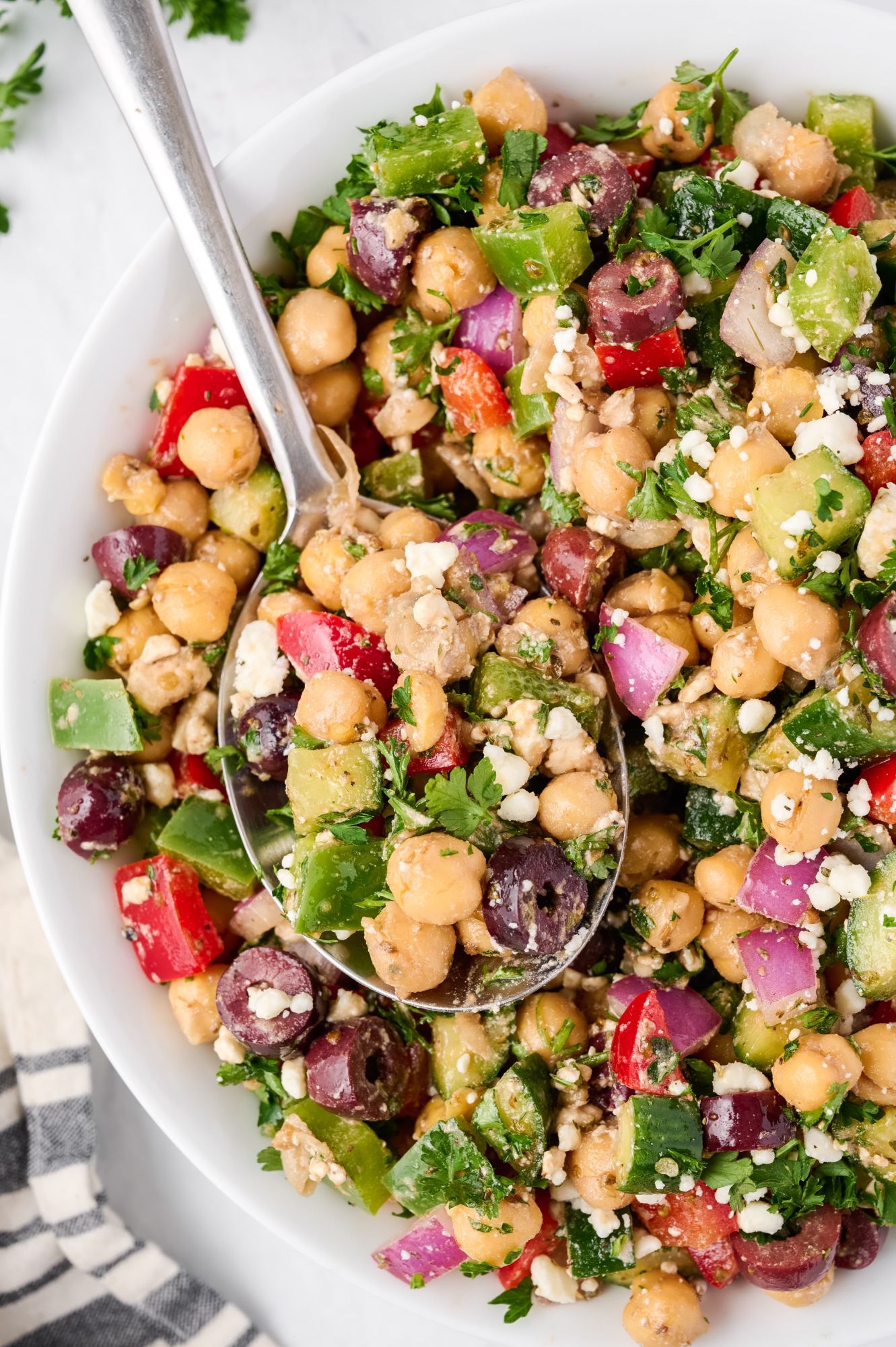 Chickpea Mediterranean salad with fresh bell peppers, cucumbers, feta cheese, herbs, and olives in a large bowl with a spoon.