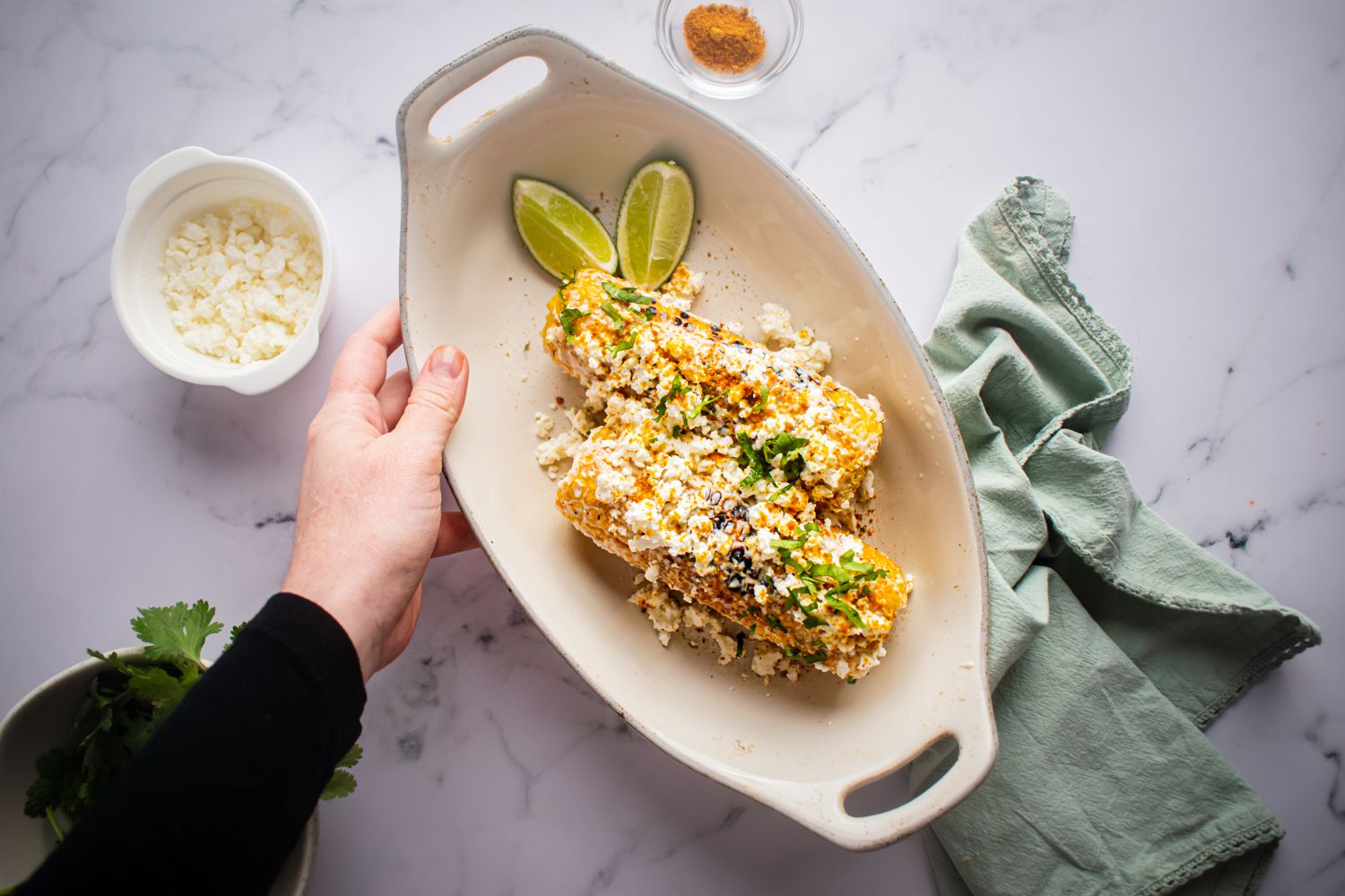 Grilled corn on the cob with lime juice, mayo, and cheese in a dish being held by a hand.