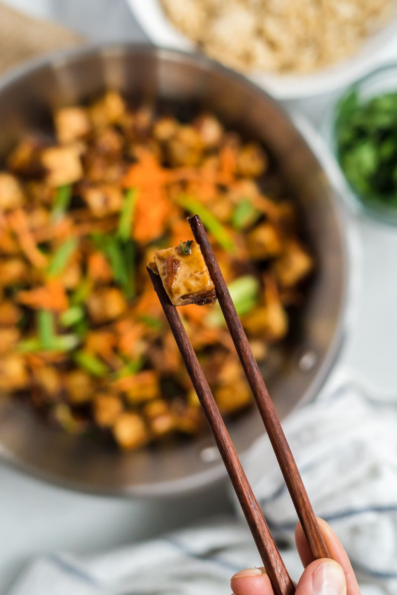 Tofu cube coated in Mongolian sauce being held above a pan with chopsticks.