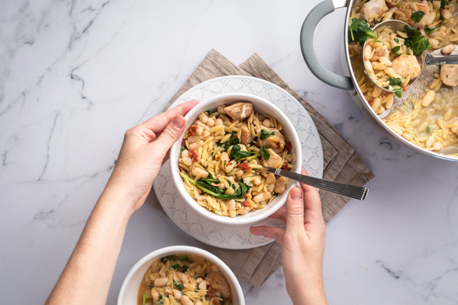 One pot orzo cooked with chicken, sundried tomatoes, spinach, and Parmesan cheese in a bowl being held by two hands.