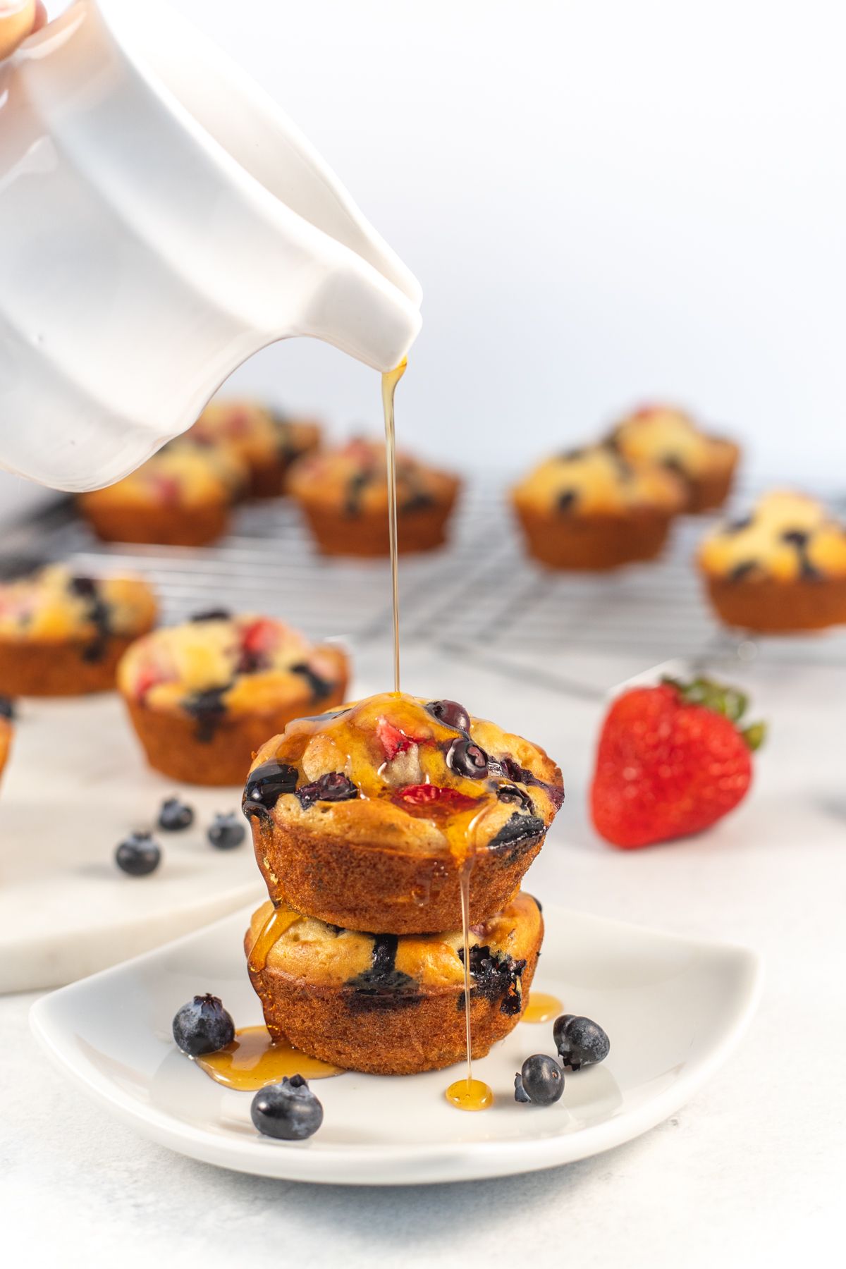 Maple syrup being poured over pancake muffins with strawberries, bananas, and blueberries.