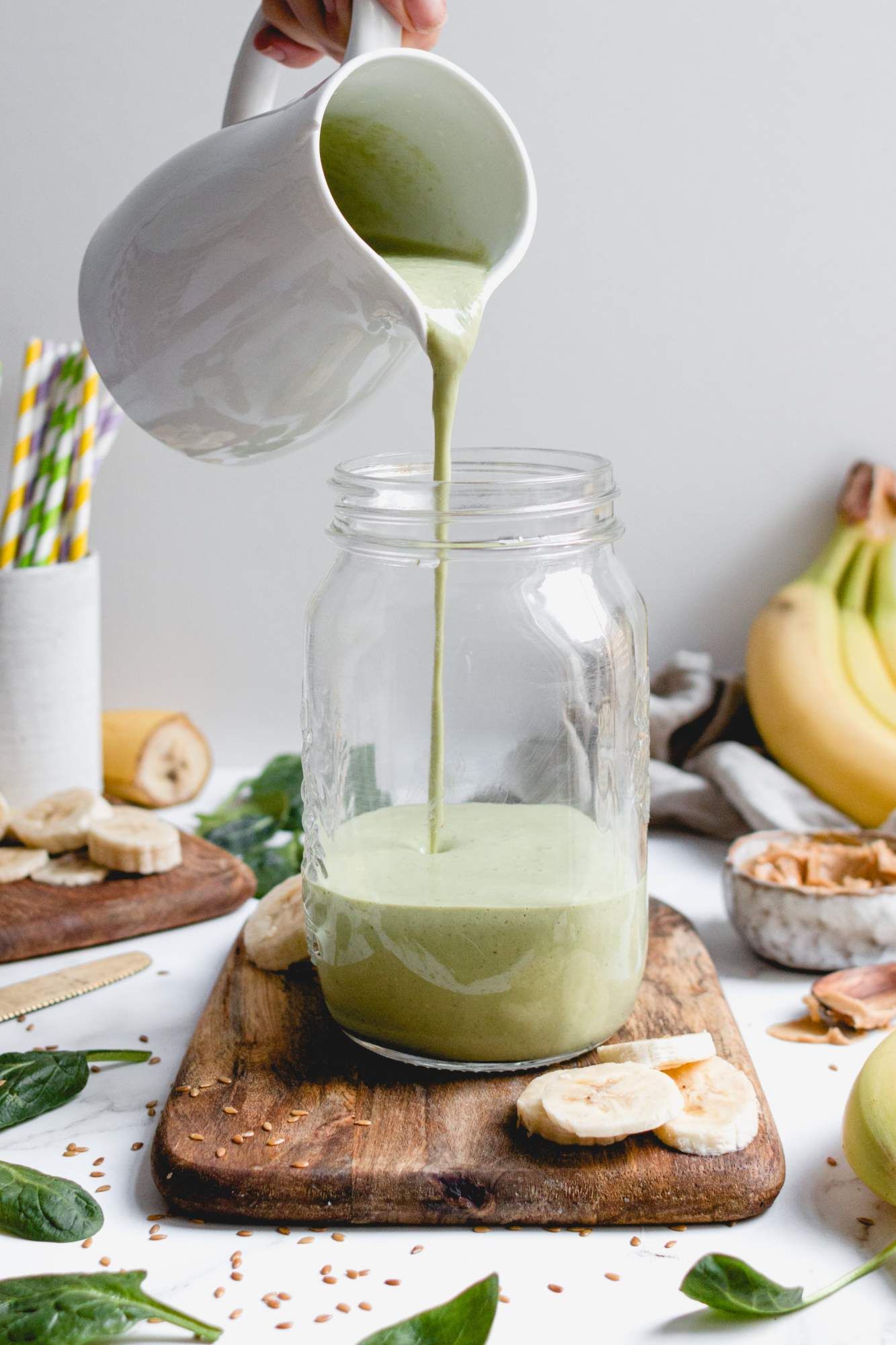 Banana green smoothie being poured into a glass with bananas, spinach, and flaxseeds on the side.