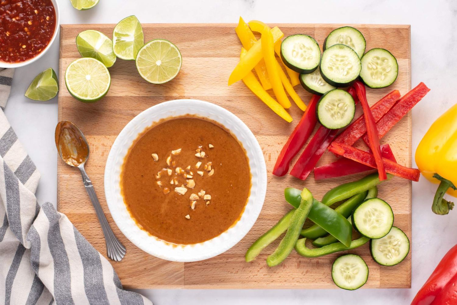 Creamy homemade peanut sauce in a small bowl with peppers, cucumbers, limes, and ginger on the side.