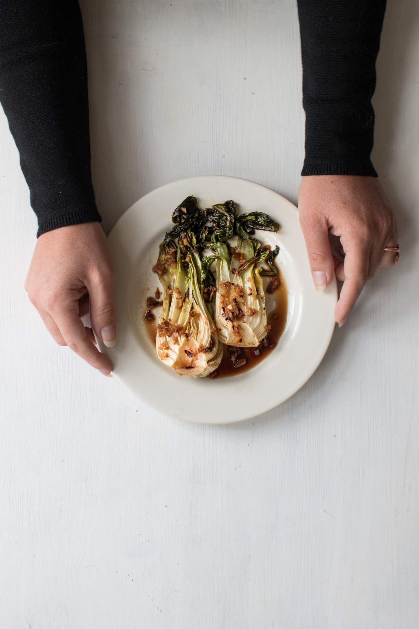 Roasted bok choy with garlic and red pepper flakes on a plate with two hands holding it.