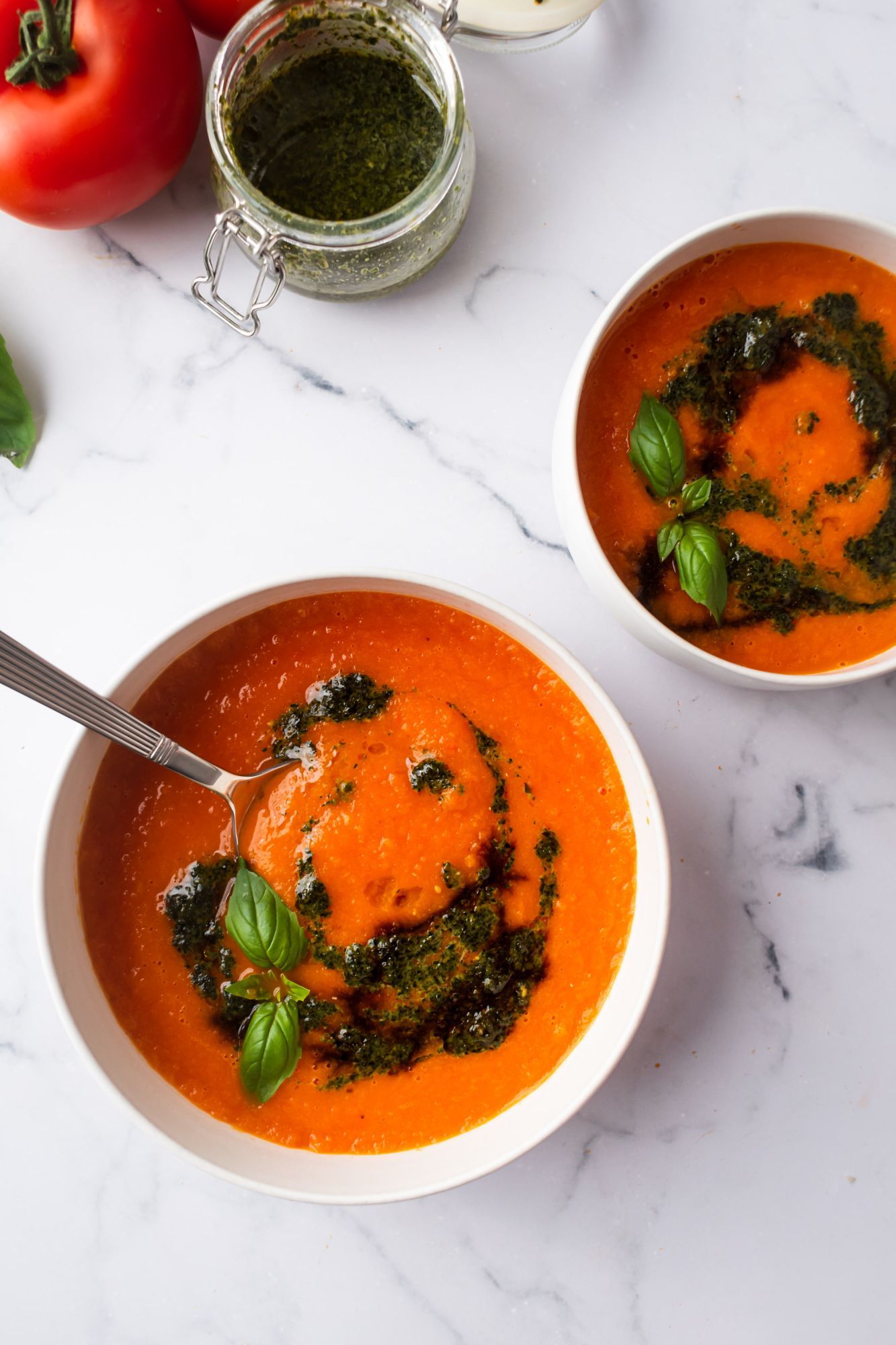 Tomato soup with basil and pesto in two bowls with fresh tomatoes on the side.