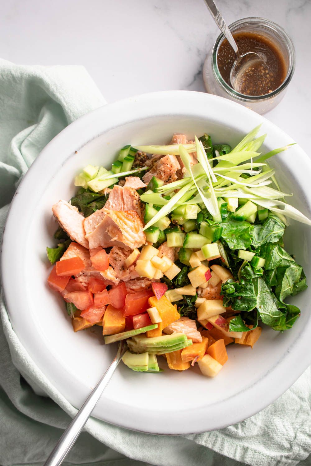 Kale salmon salad with sweet potatoes, tomatoes, apples, and avocado in a bowl with dressing on the side.