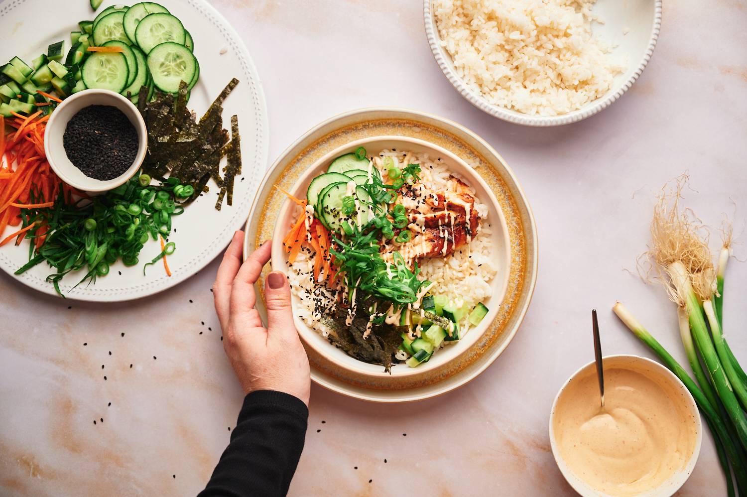 Deconstructed salmon sushi roll bowls with seaweed, cucumbers, carrots, green onions, and sushi rice in a bowl with a hand.