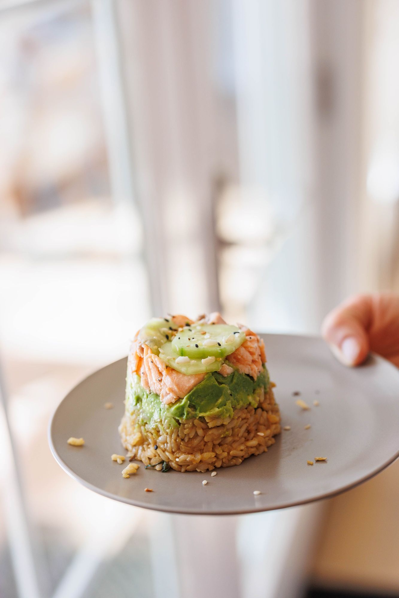 Sushi stack with brown rice, cooked salmon, cucumbers, avocado, and sesame seeds on a gray plate.