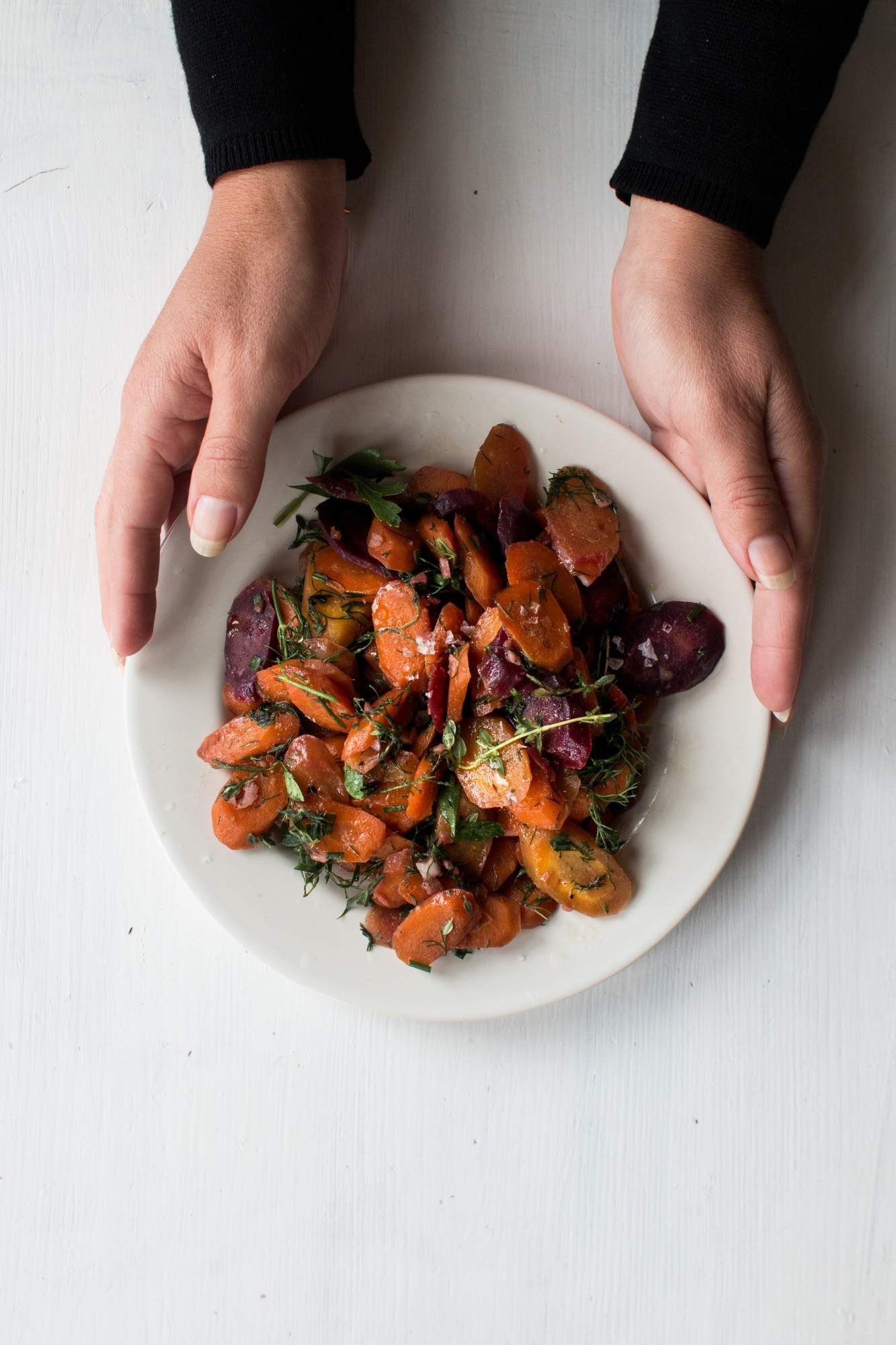 Carrots that are sautéed in a bowl with fresh herbs being held by two hands. in a 