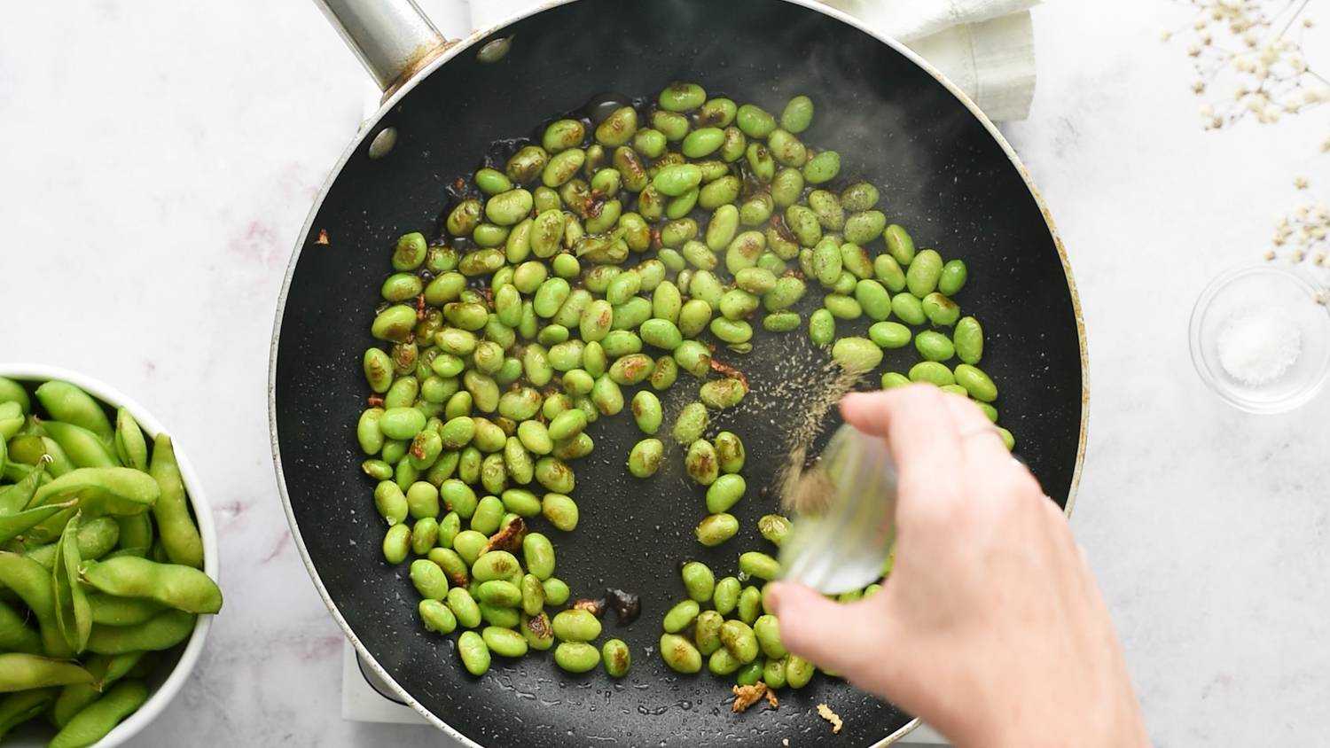 Shelled edamame cooking in a skillet with garlic, black pepper, soy sauce, and sesame oil.